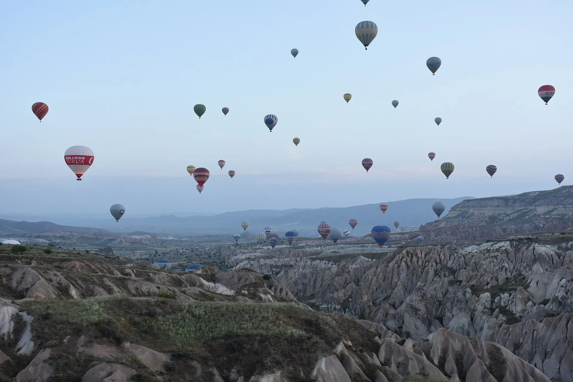 Dutzende Heißluftballons in der Morgendämmerung über den Tälern bei Göreme, unten die hellen, zerfurchten Tuffgrate