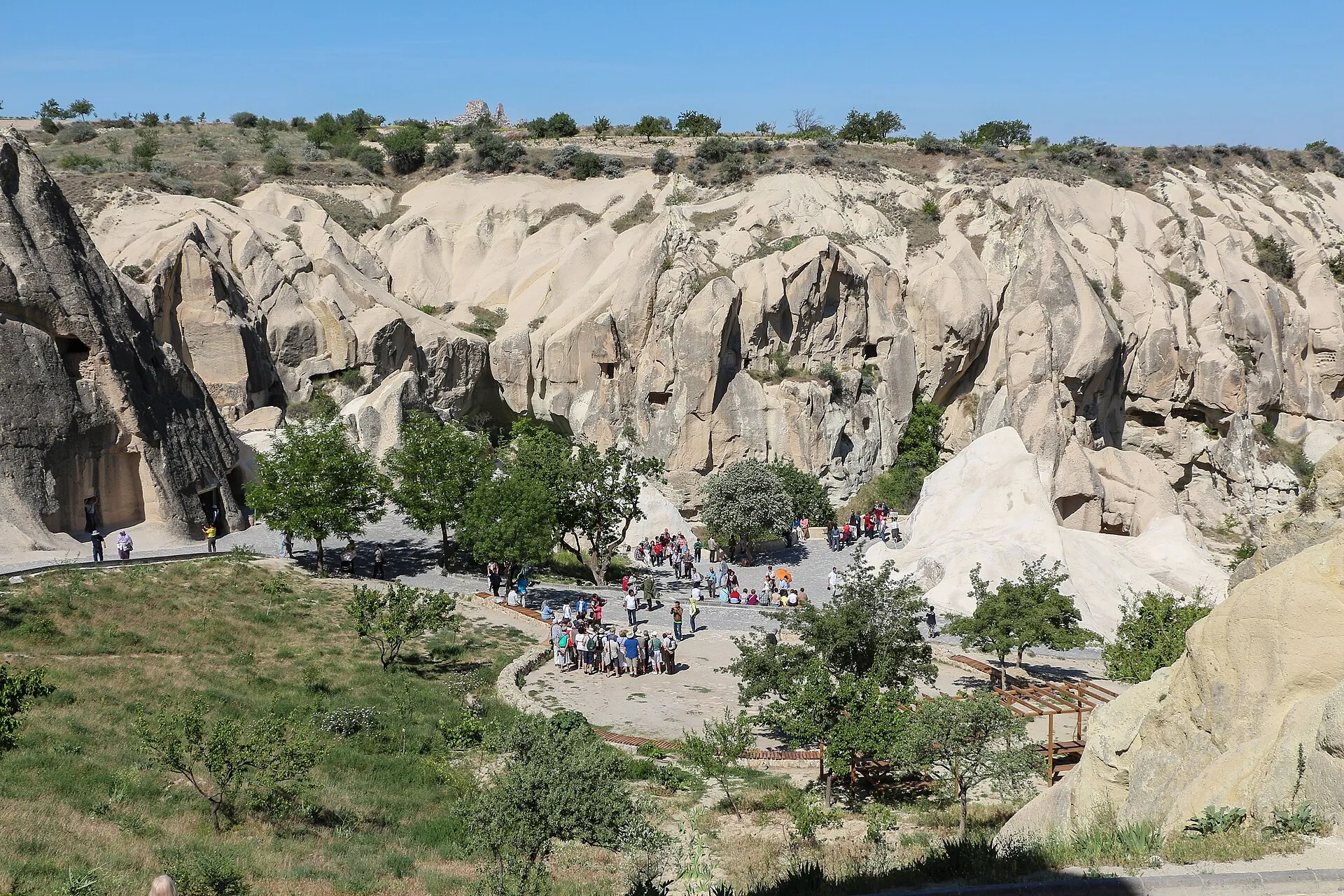 Das Freilichtmuseum Göreme von oben: helle Tuffwände mit den Eingängen der Felsenkirchen, davor Reisegruppen auf dem Vorplatz zwischen Bäumen