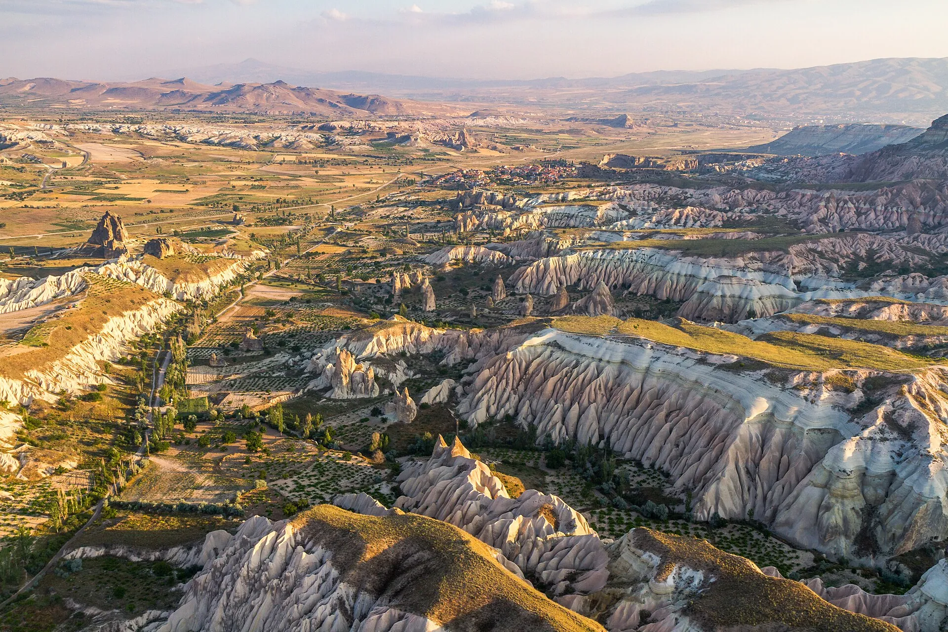 Blick aus der Luft über das Rosental und den Ort Çavuşin bei Göreme: rosa und weiße Tuffkämme, dazwischen Felder und Bäume, am Horizont die Berge