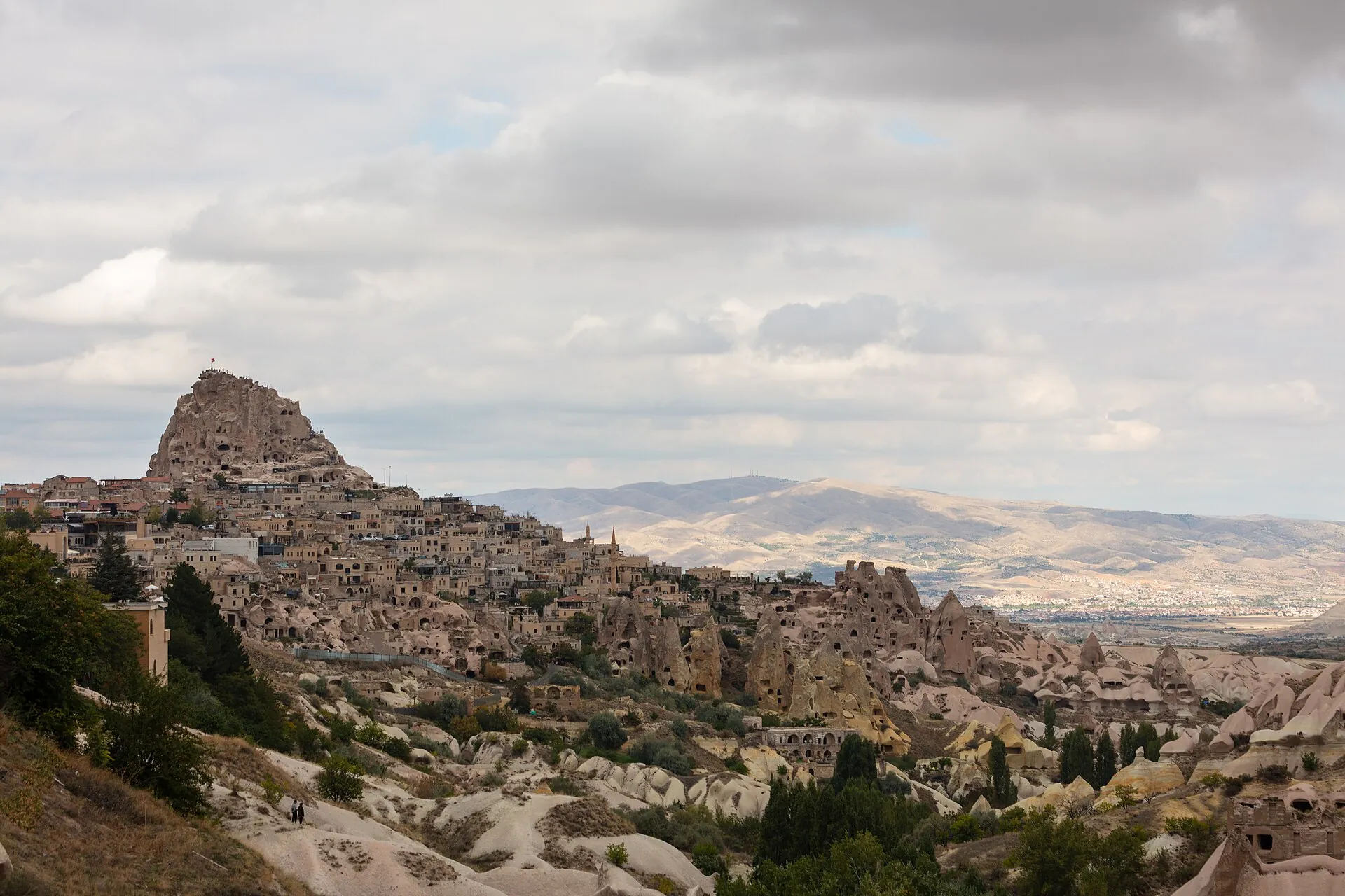 Blick über Uçhisar in Kappadokien: der Burgfelsen über dem Ort, davor Höhlenhäuser und Feenkamine, dahinter die Ebene bis zum Horizont