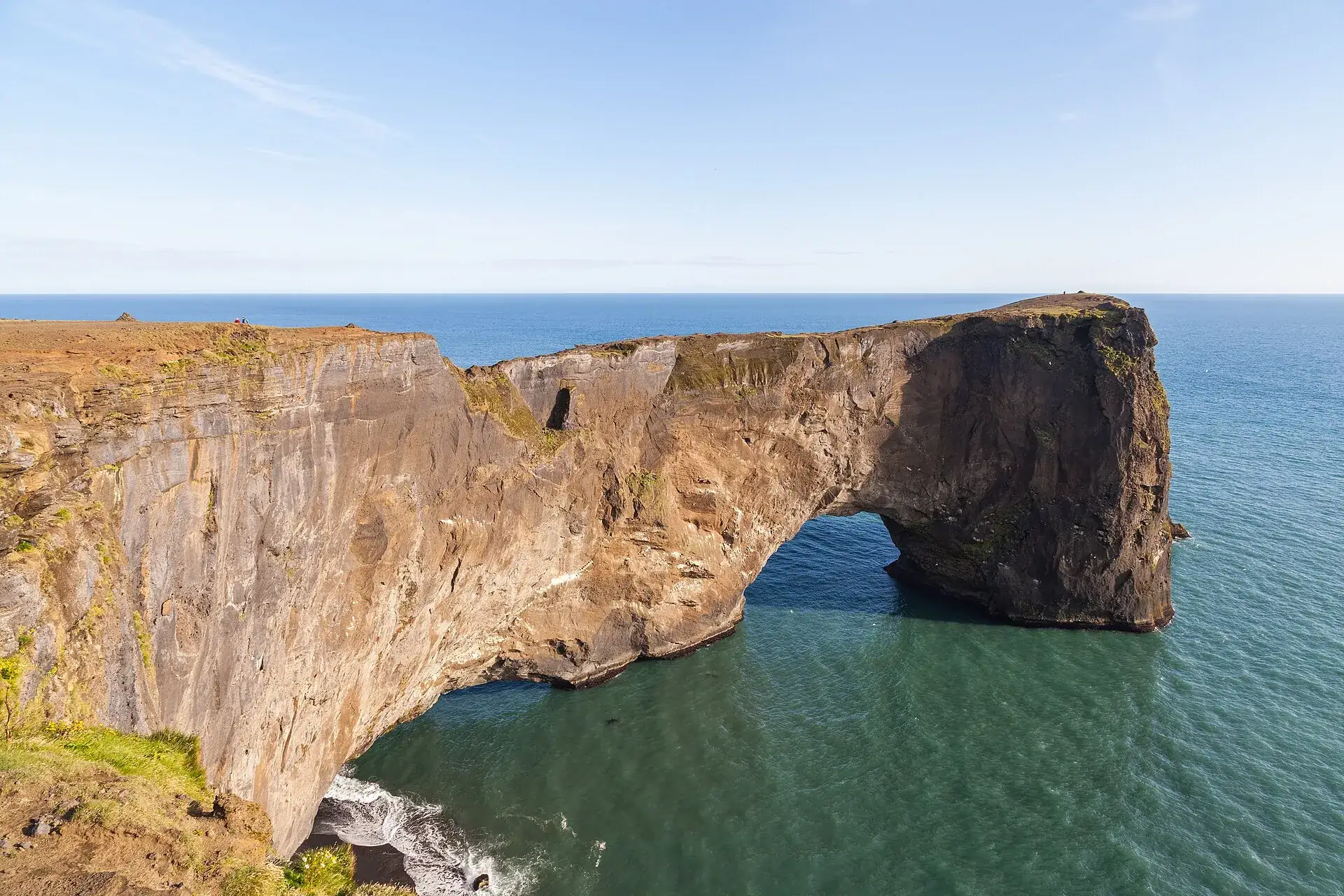 The rock arch of Dyrhólaey juts into the sea, green water below, blue sky