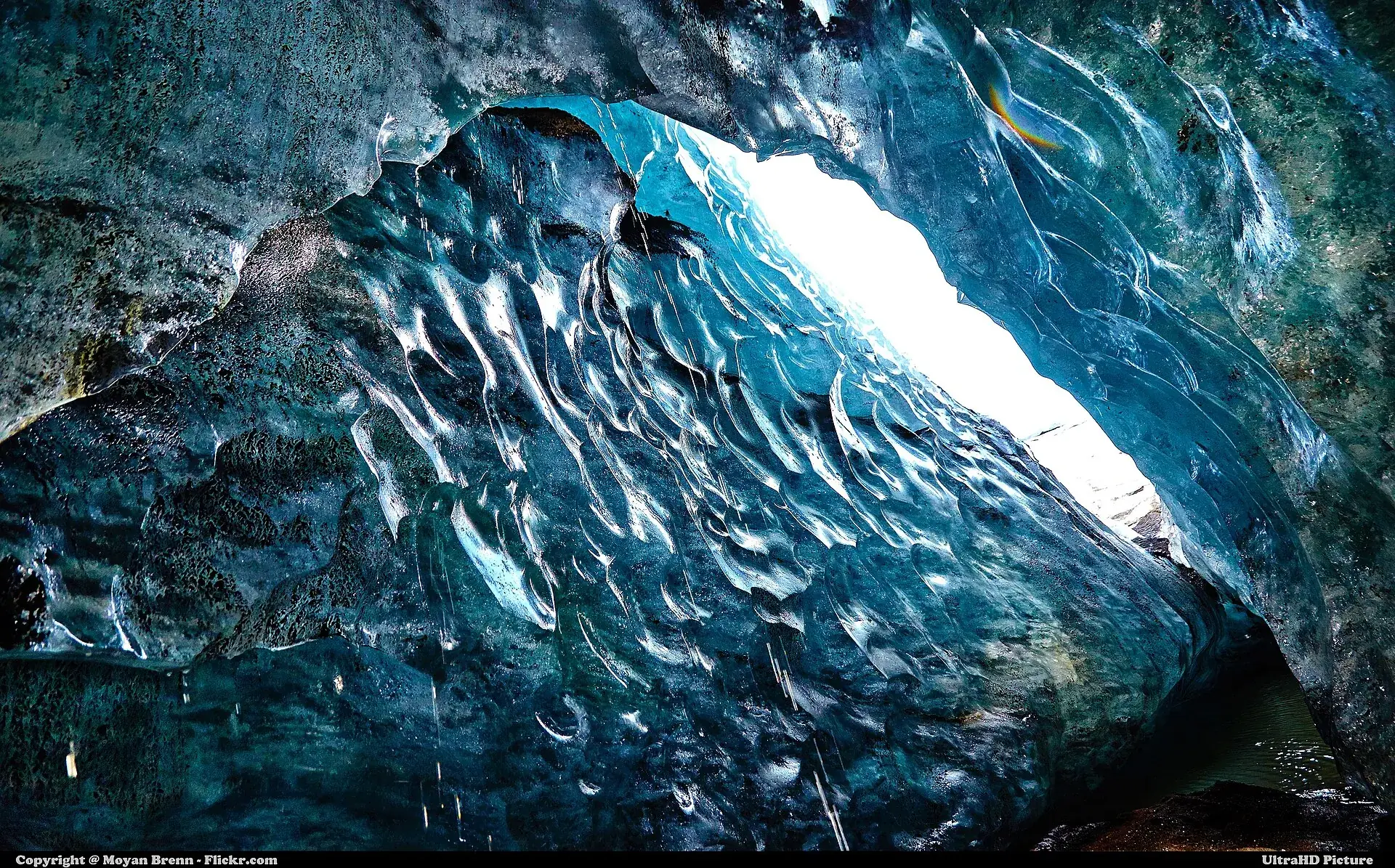 Looking out of an ice cave at Mýrdalsjökull: blue ice with dark ash bands, daylight falling through the opening