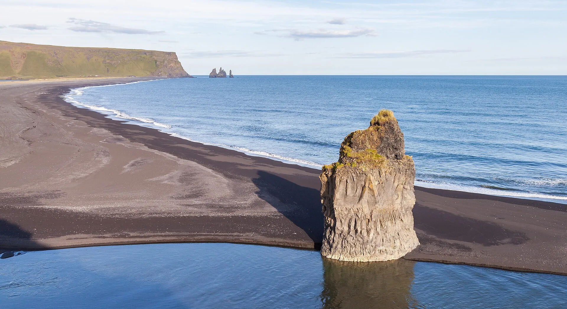 The black beach of Reynisfjara from above: surf, a single rock tower, Dyrhólaey far in the background