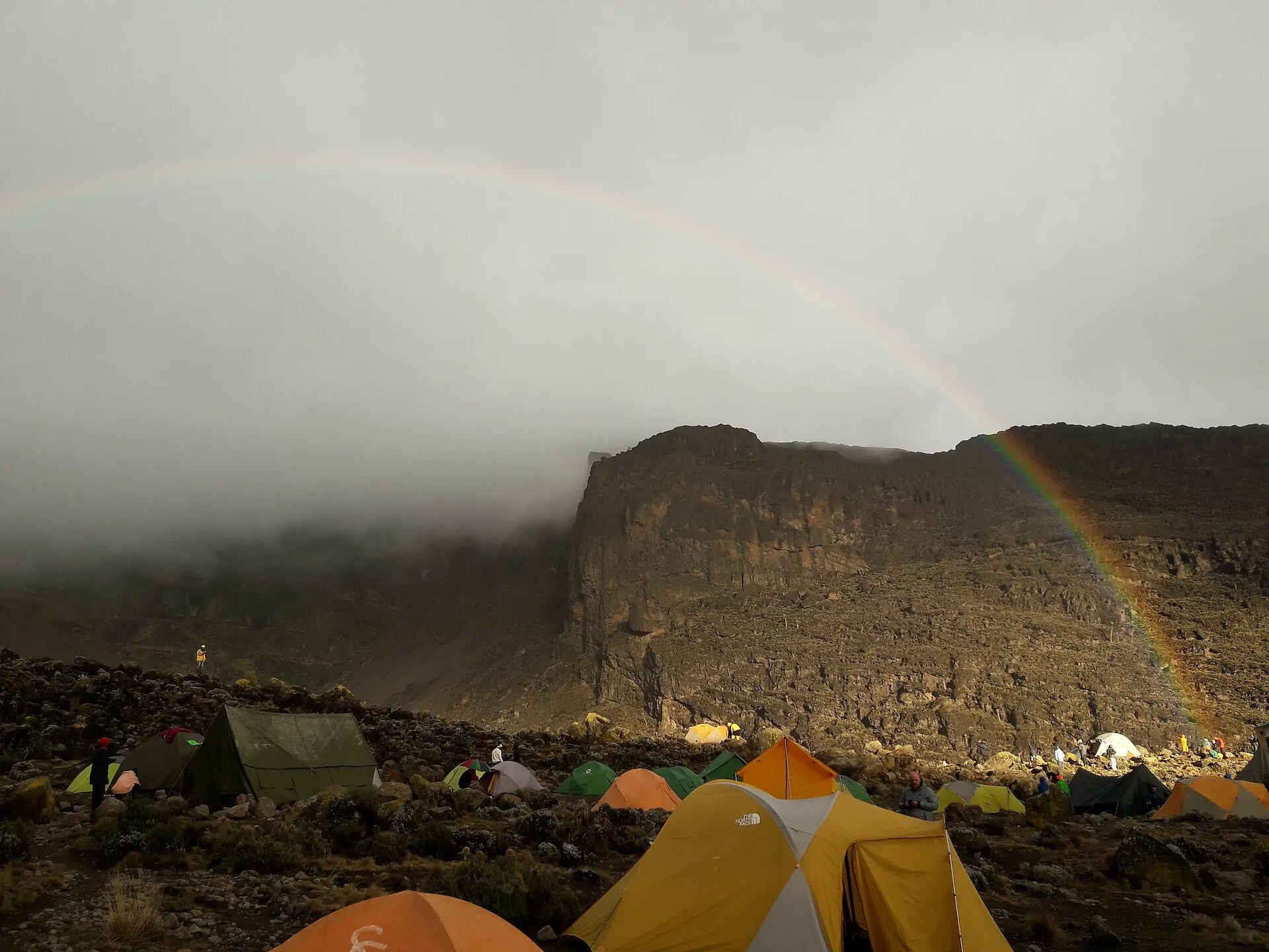 Tents at Barranco Camp on Kilimanjaro under a rainbow, the dark rock wall behind them in cloud