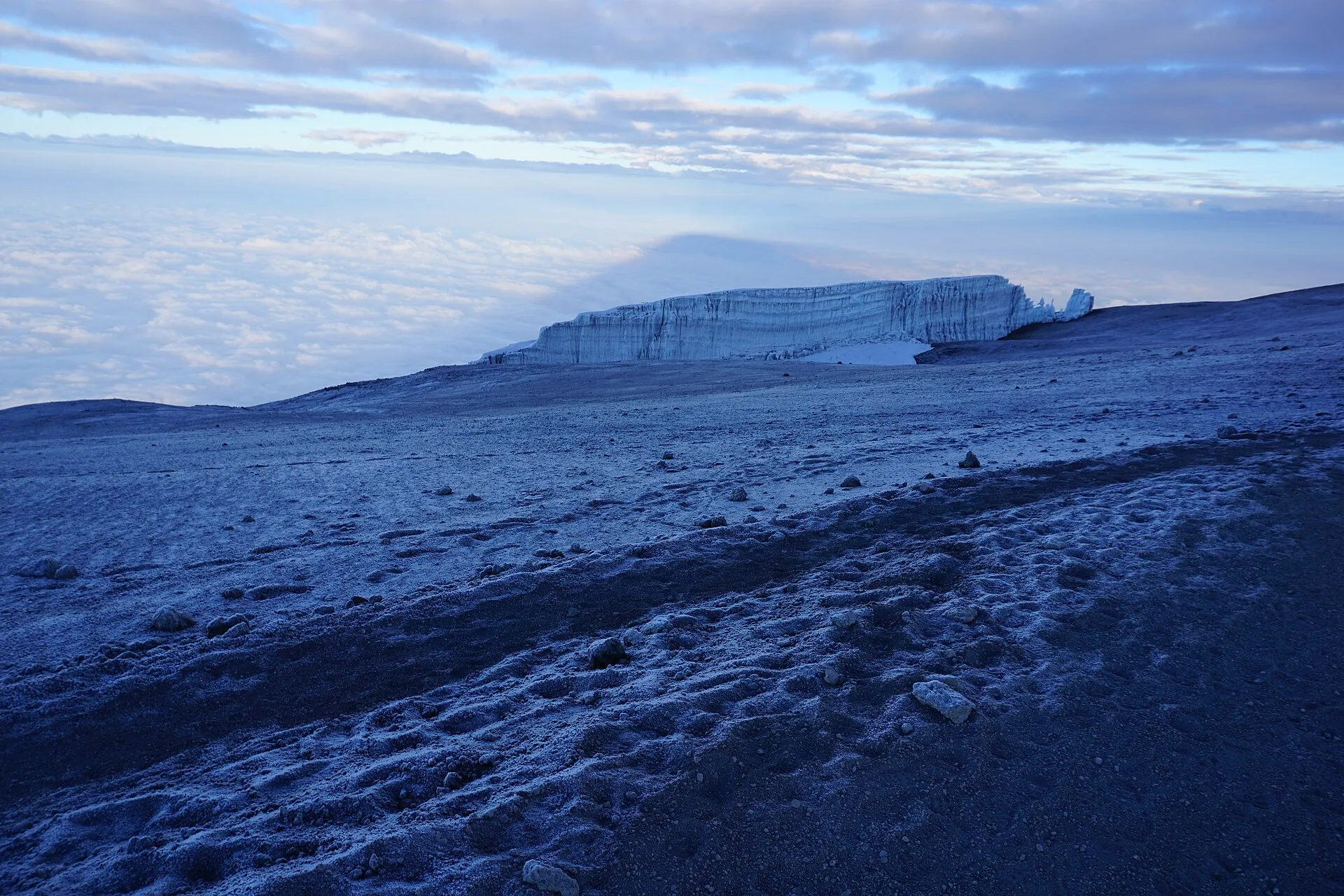 The edge of a glacier near the summit of Kilimanjaro at dawn, frozen scree in front and a sea of cloud below