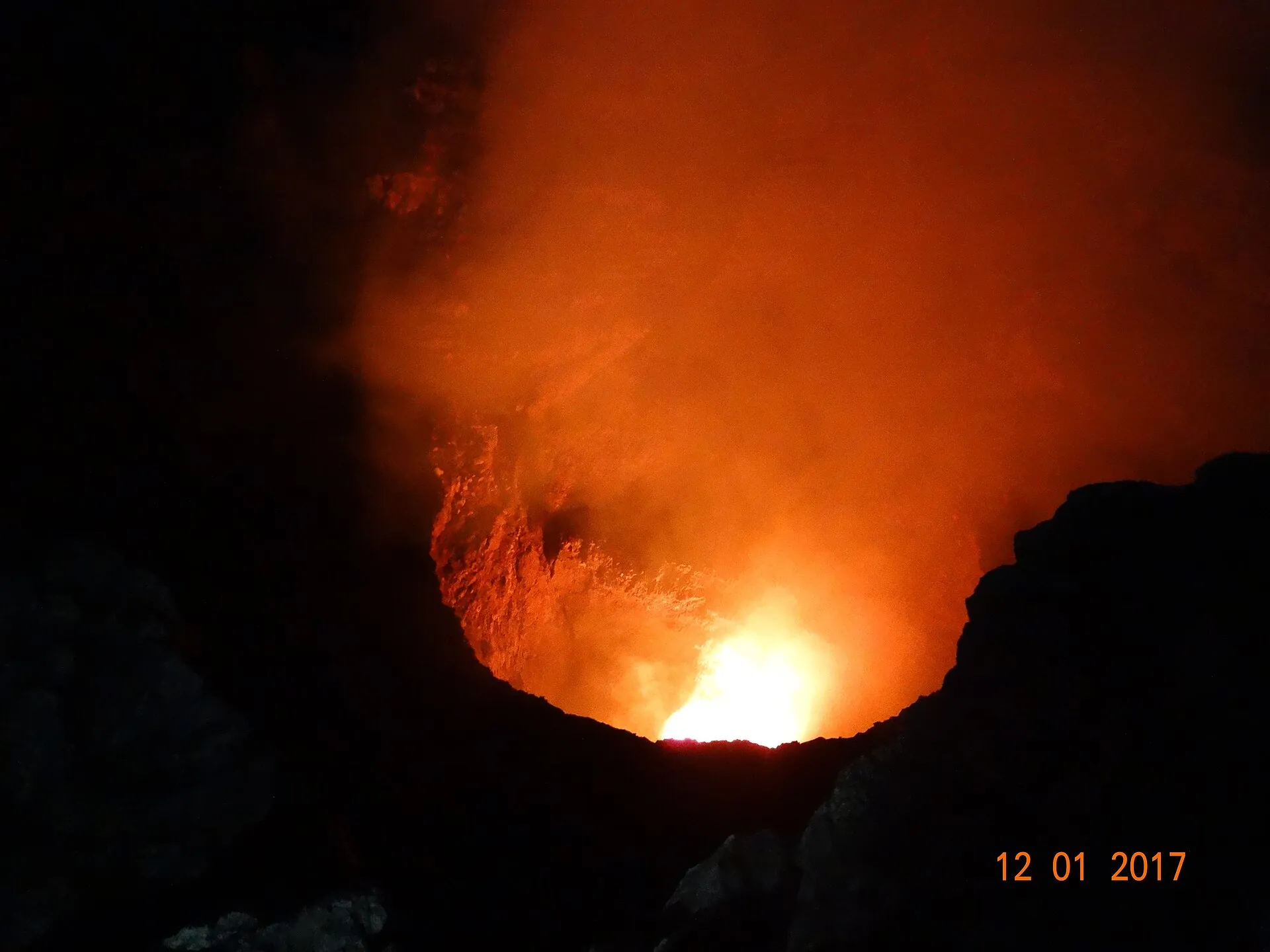 Glühende Lava im Cráter Santiago des Masaya bei Nacht, Januar 2017: orangefarbene Glut hinter Gasschwaden, schwarze Kraterwände
