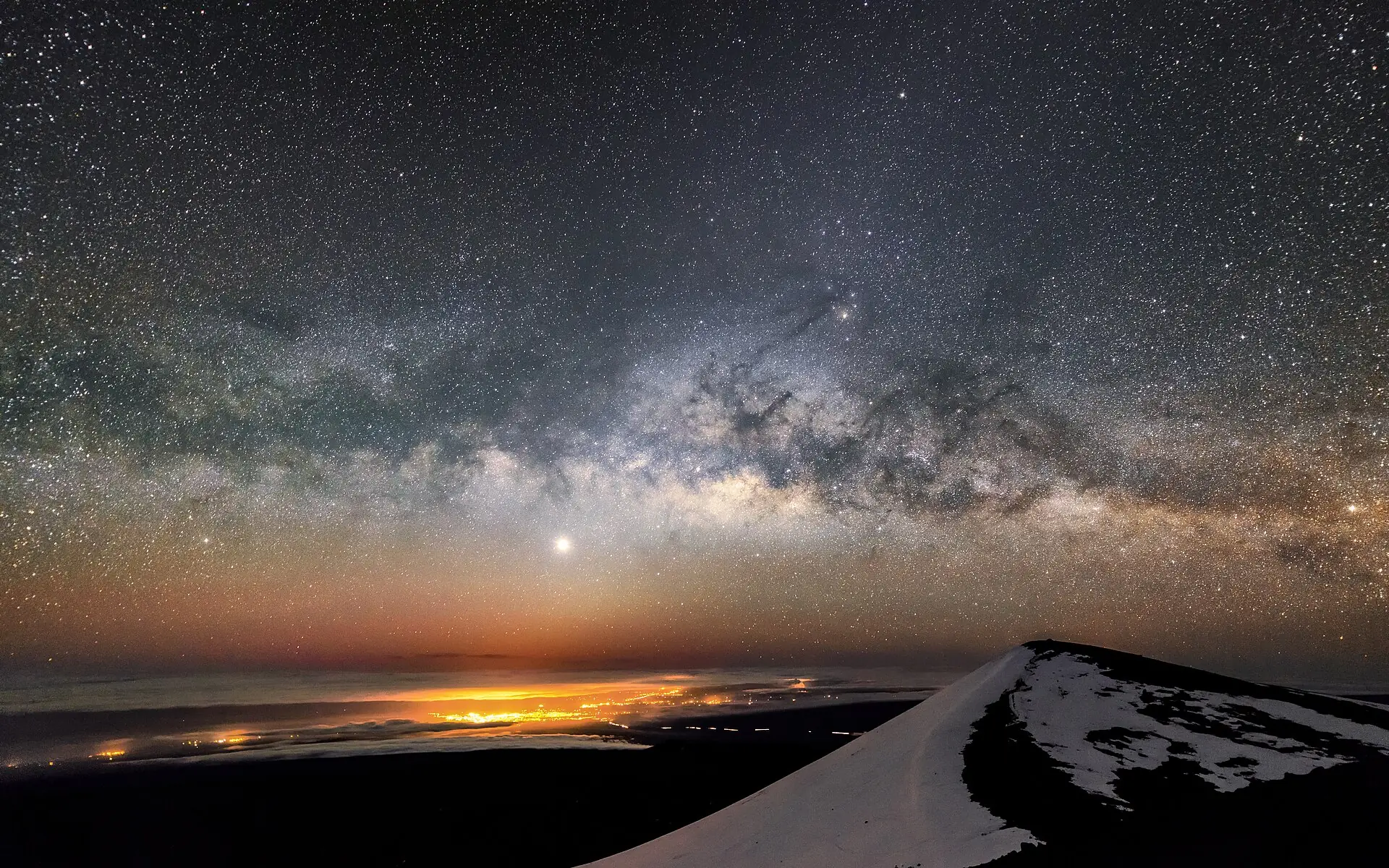 The Milky Way and Jupiter over Mauna Kea at night, the lights of Hilo below and a snow-covered cinder cone in the foreground