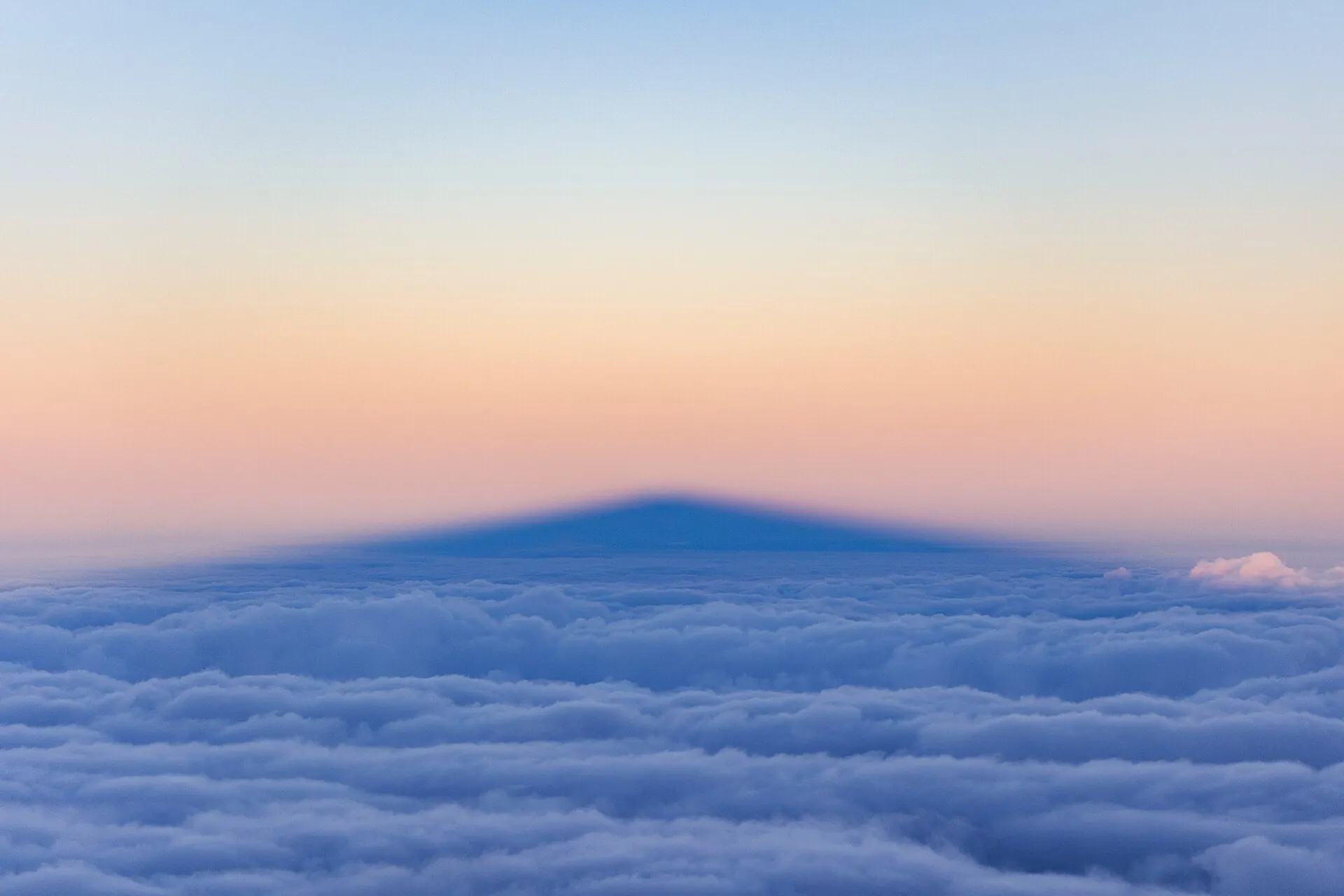 The shadow of Mauna Kea falls as a dark triangle on the unbroken cloud deck below the summit, under a pastel sky