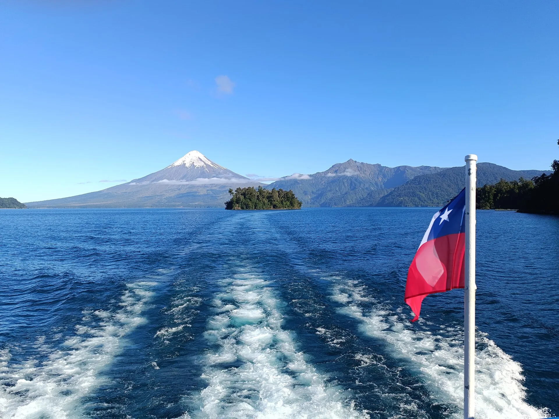 The wake of an excursion boat on the deep blue Lake Todos los Santos, a Chilean flag at the stern, Osorno in the background