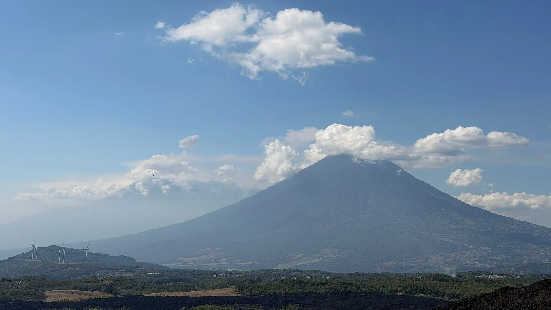 View from Pacaya across dark lava fields and green highlands to the cone of Volcán de Agua under fair-weather clouds