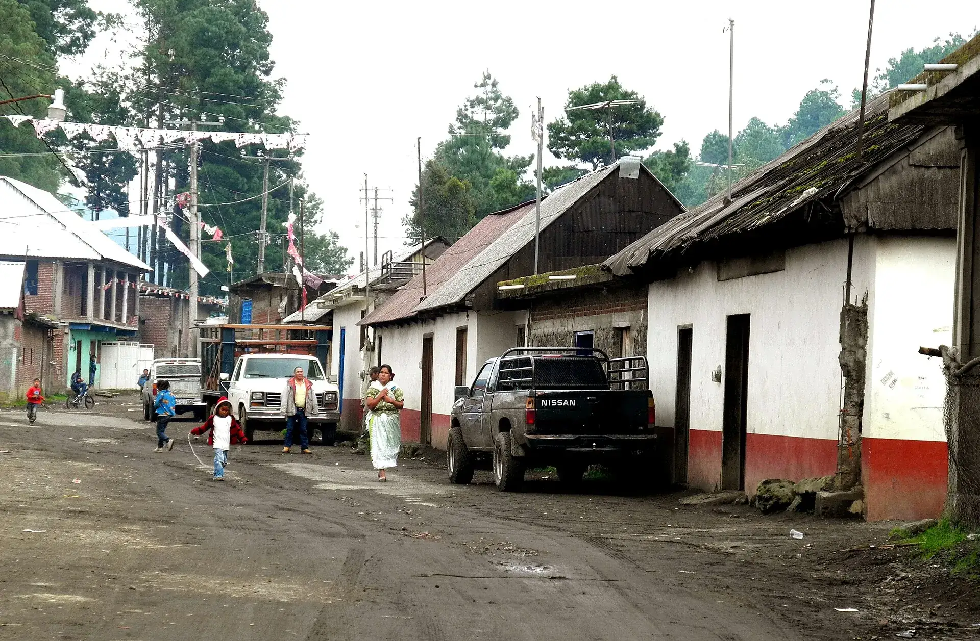 Eine Straße in Angahuan, Michoacán: weiße Häuser mit Holzdächern, Menschen und ein Pickup, dahinter Kiefern