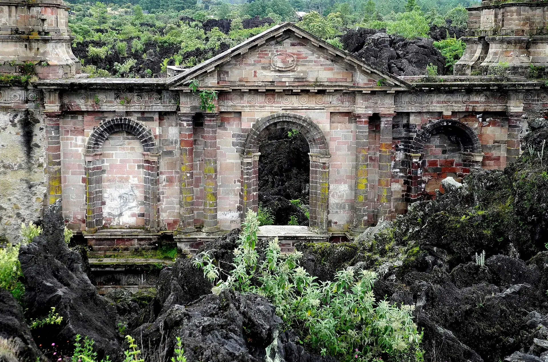 Die Fassade der Kirchenruine von San Juan Parangaricutiro mit drei Bögen und Flechten auf dem Stein, davor Lavablöcke und Farne