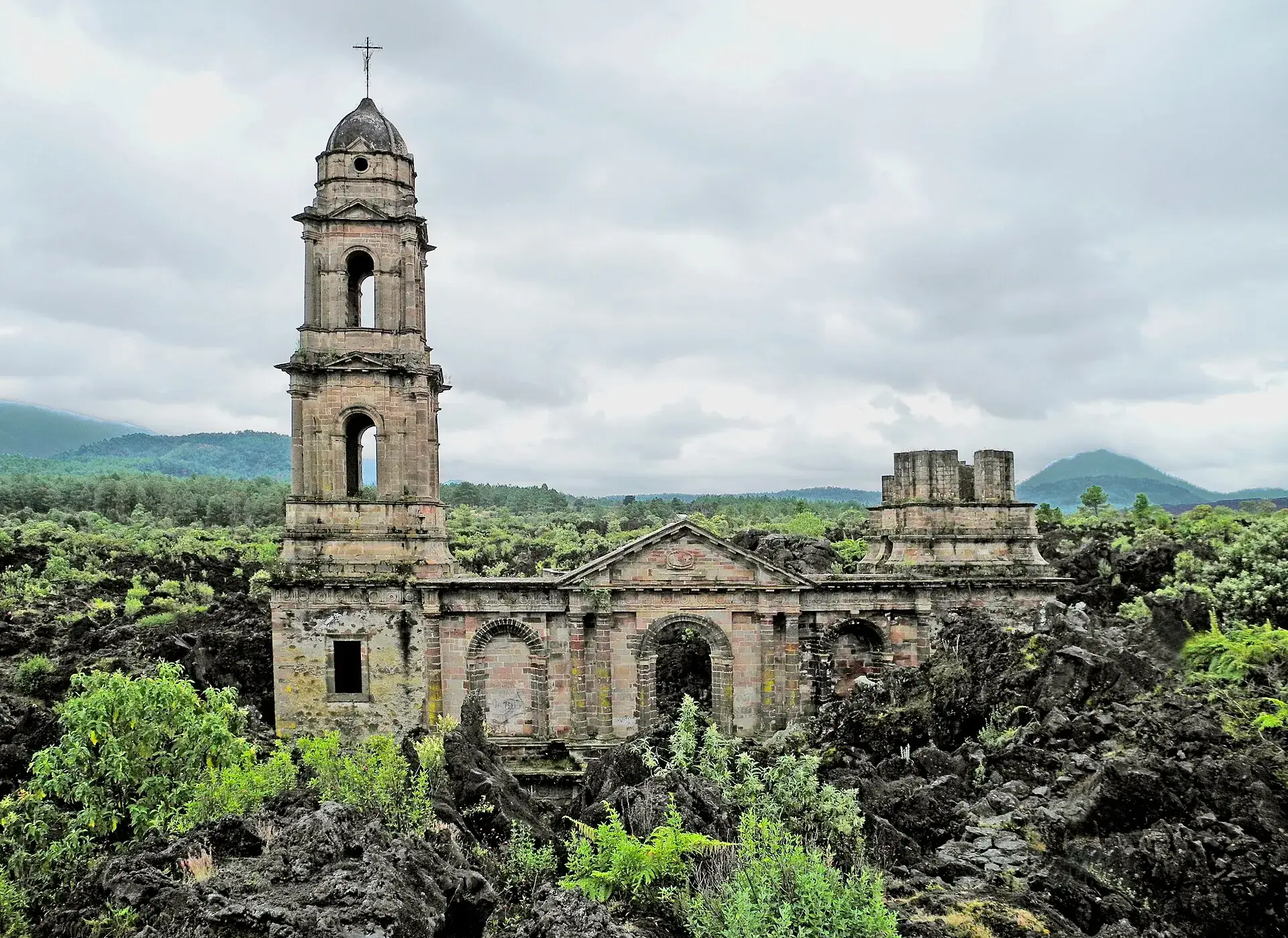 Die beiden Türme der Kirchenruine von San Juan Parangaricutiro ragen aus dem schwarzen Lavafeld, dahinter Wald und Hügel unter bedecktem Himmel