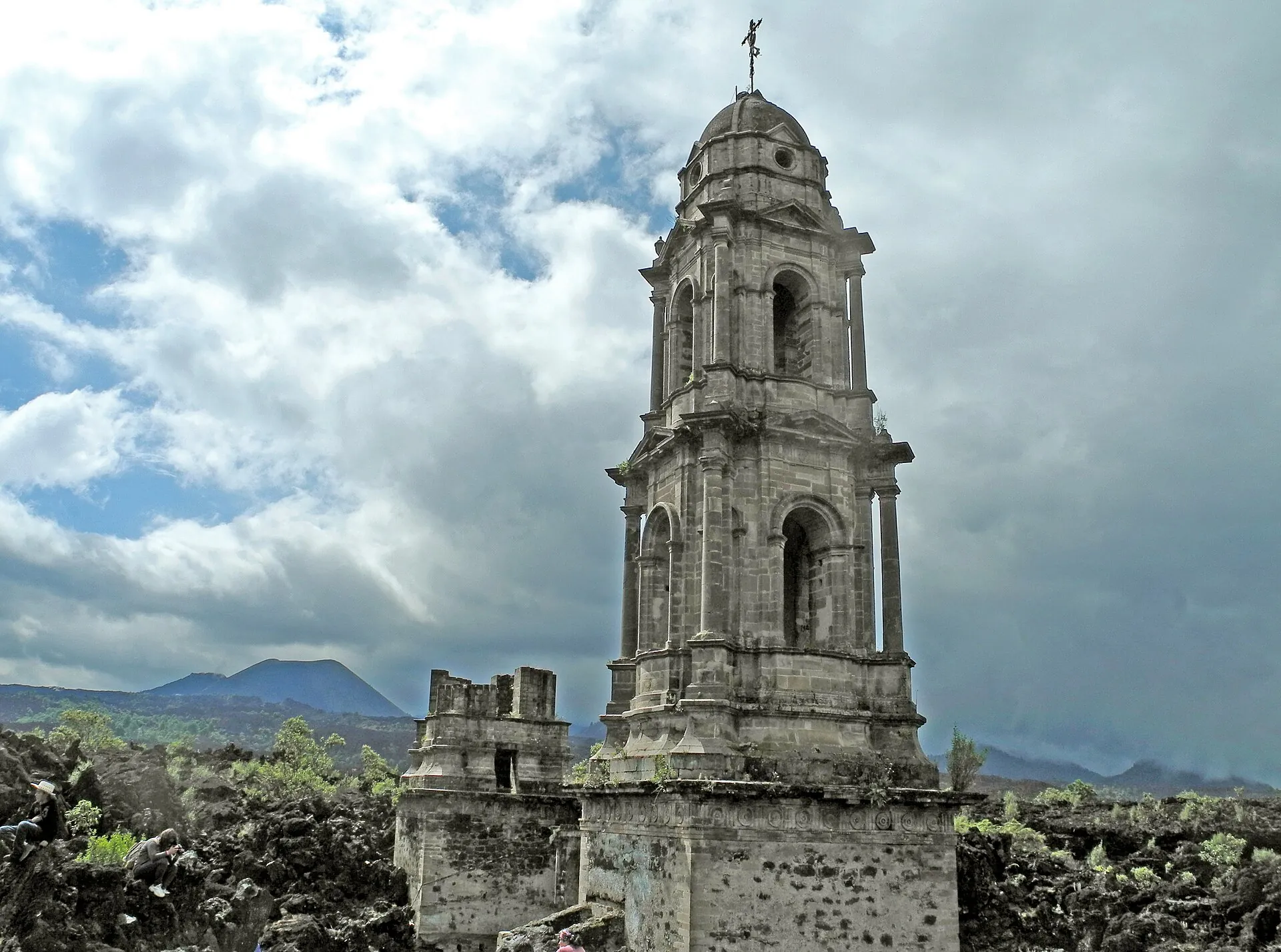 Der Kirchturm von San Juan Parangaricutiro ragt aus schwarzer Lava, darüber ein dramatischer Wolkenhimmel, im Hintergrund der Kegel des Paricutín