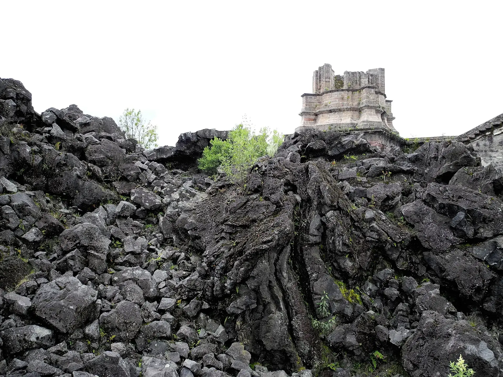 Der obere Teil eines Kirchturms von San Juan Parangaricutiro ragt aus einem Wall schwarzer Blocklava, dazwischen junge Bäume