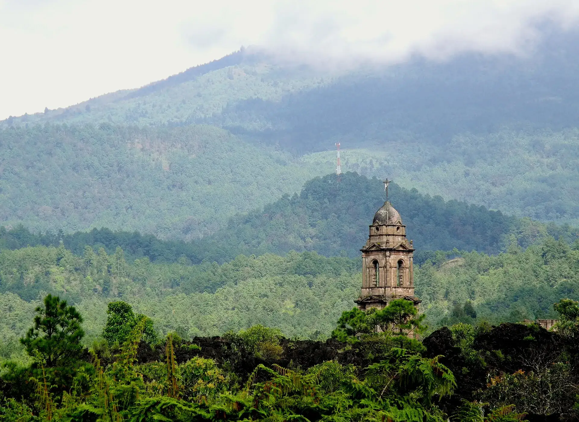 Der Kirchturm von San Juan Parangaricutiro ragt über den Wald, dahinter bewaldete Hügel und tief hängende Wolken