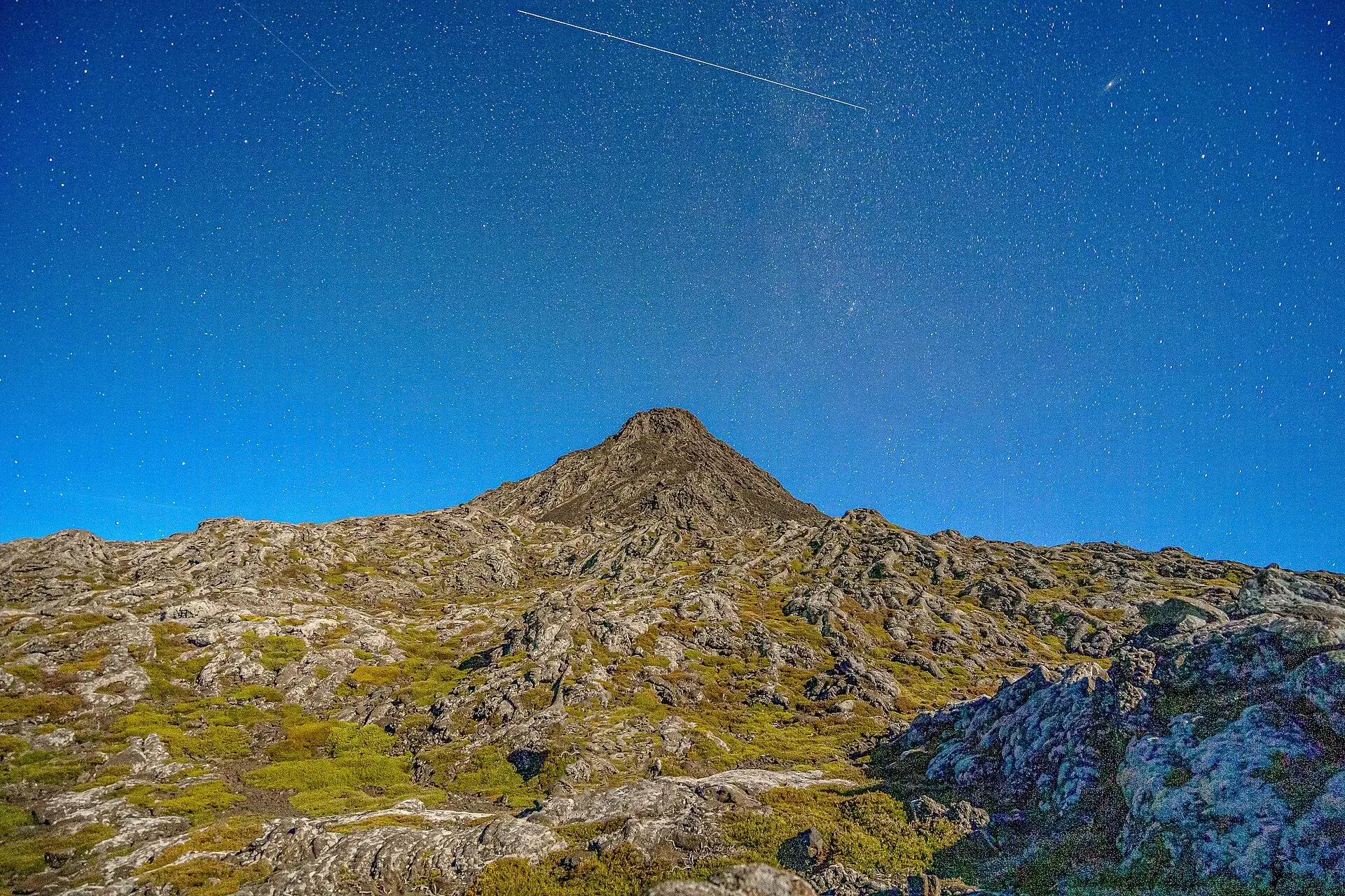 Der Pico bei Nacht unter einem Sternenhimmel, im Vordergrund Lavafelder mit Flechten, am Himmel die Spur eines Kometen