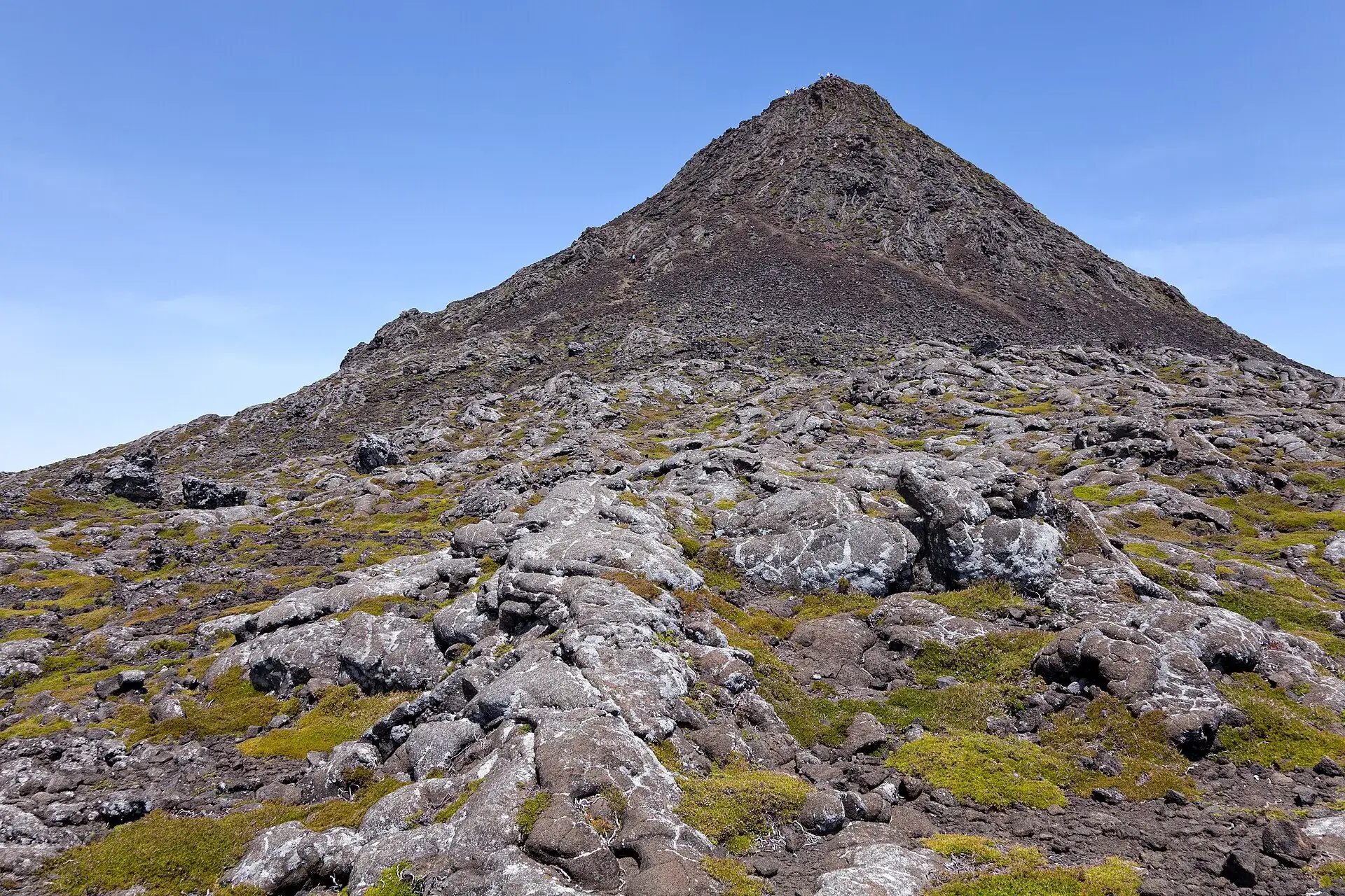 Der Piquinho, der Gipfelkegel des Pico: 70 Meter dunkle Lava vor blauem Himmel, Wanderer auf dem Kraterrand