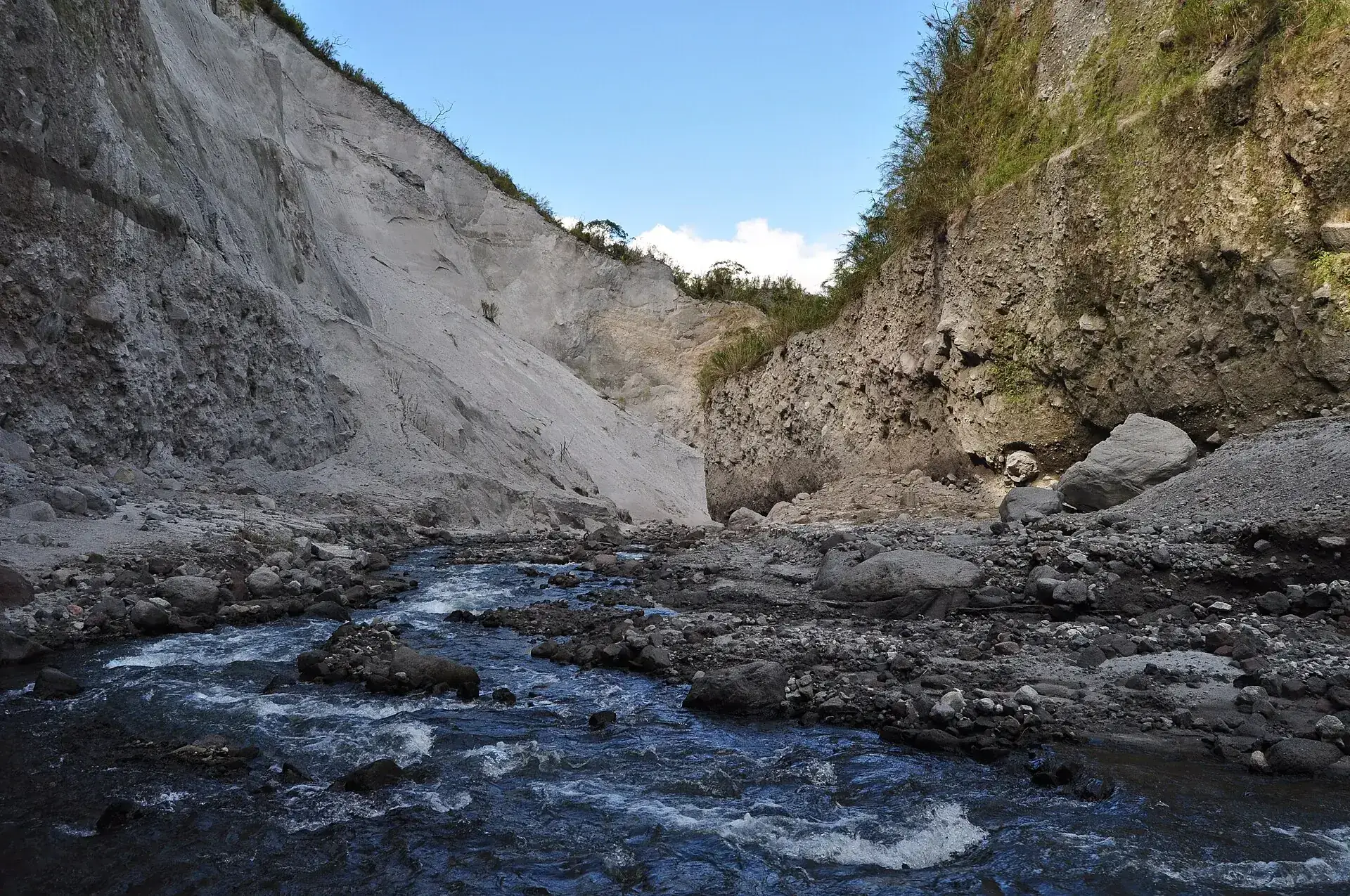 Ein klarer Bach fließt über dunkle Steine durch das Flussbett am Pinatubo, dahinter eine hohe hellgraue Aschewand