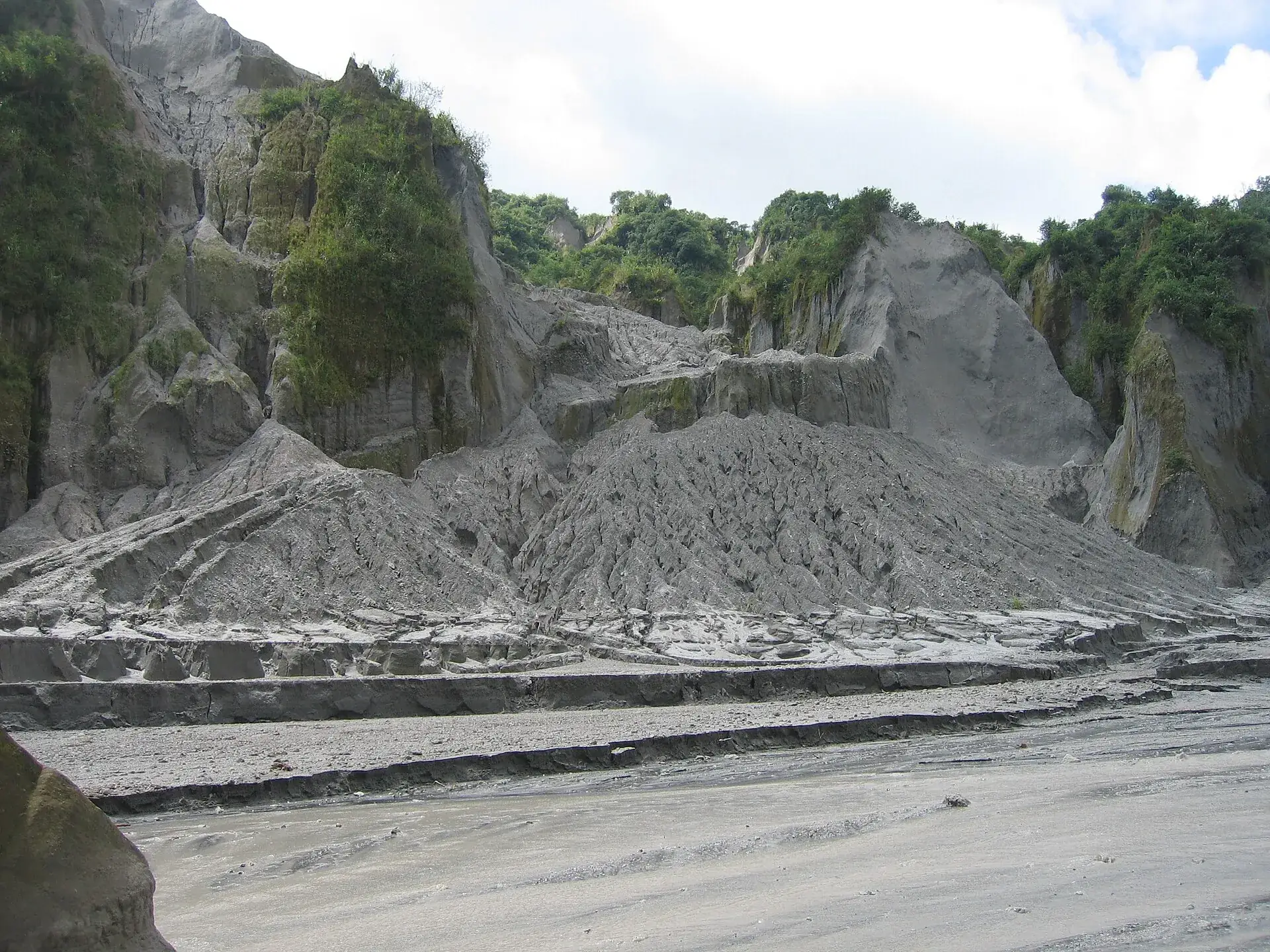Hellgraue Lahar-Ablagerungen am Pinatubo: eine ausgewaschene Aschewand mit Rinnen, davor das breite, flache Flussbett
