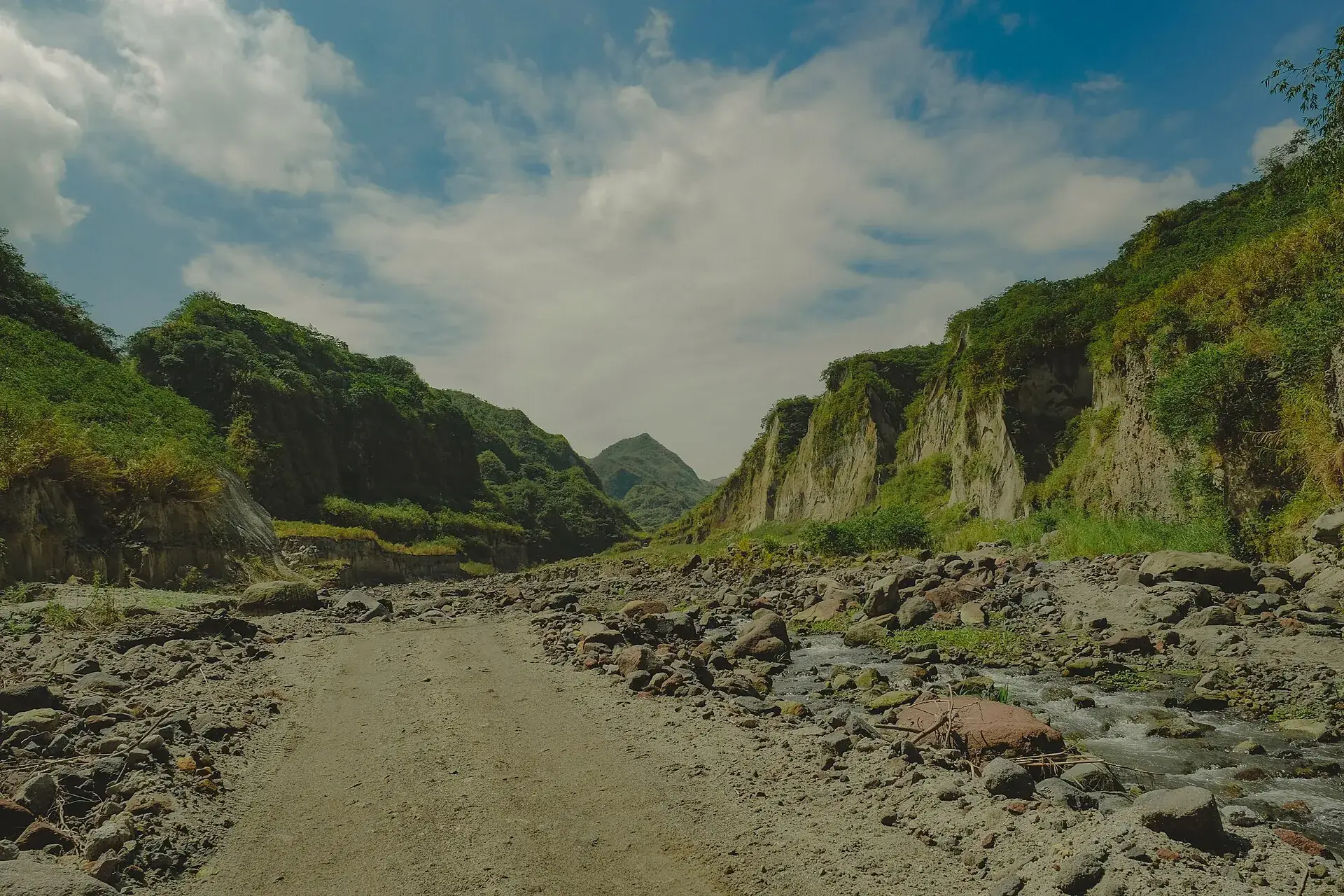 Die Jeep-Piste im Crow Valley am Pinatubo: eine steinige Fahrspur im breiten Flussbett, links und rechts hohe Aschewände unter Wolken