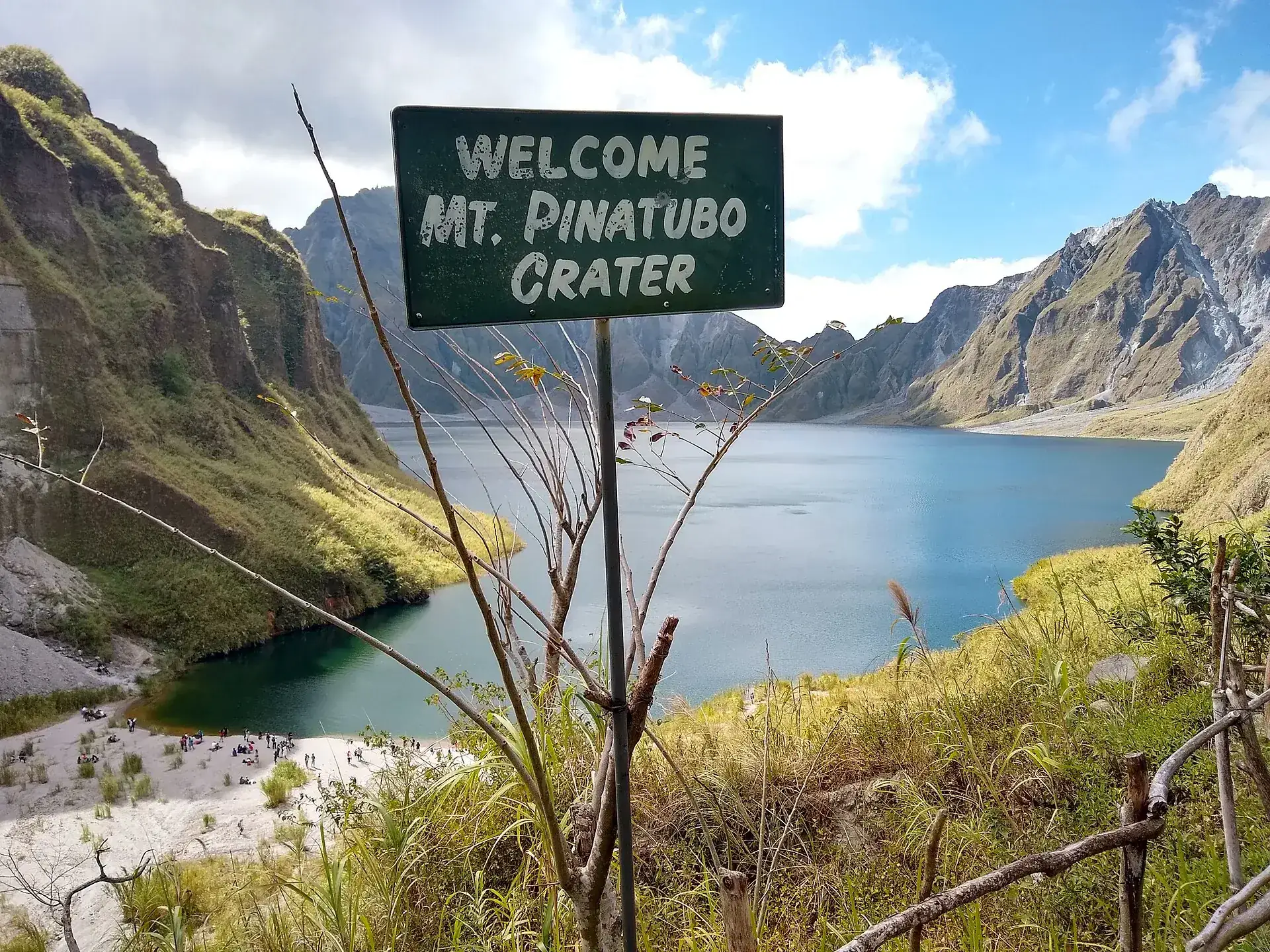 Grünes Schild „Welcome Mt. Pinatubo Crater“ am Aussichtspunkt, dahinter der blaue Kratersee und die Calderawände