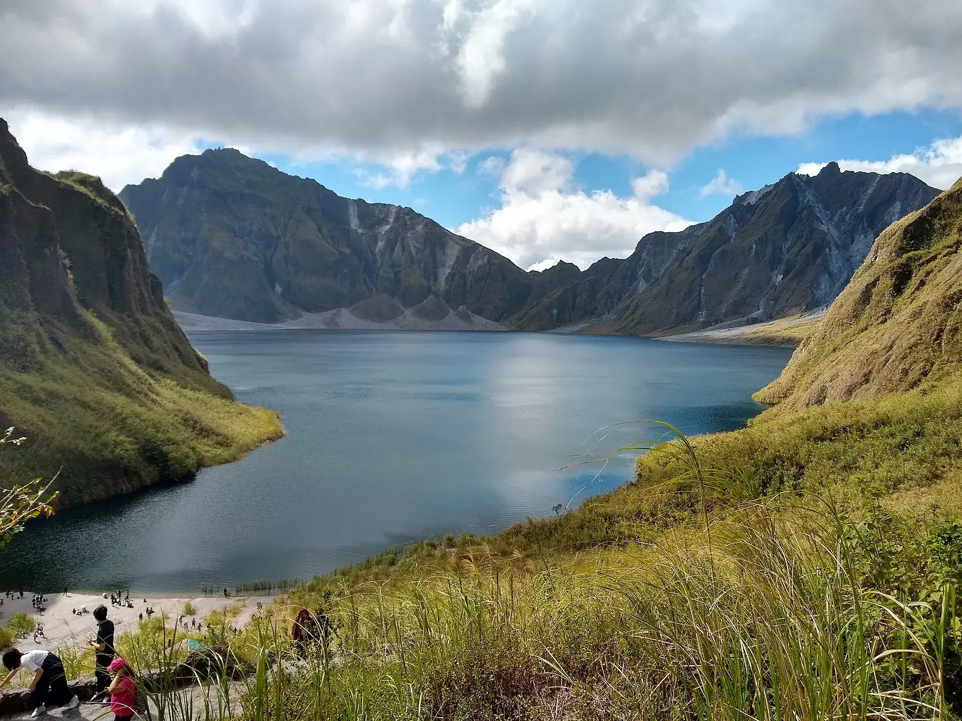 Besucher sitzen am grasbewachsenen Hang über dem Kratersee des Pinatubo, das Wasser blaugrün, gegenüber die Calderawand