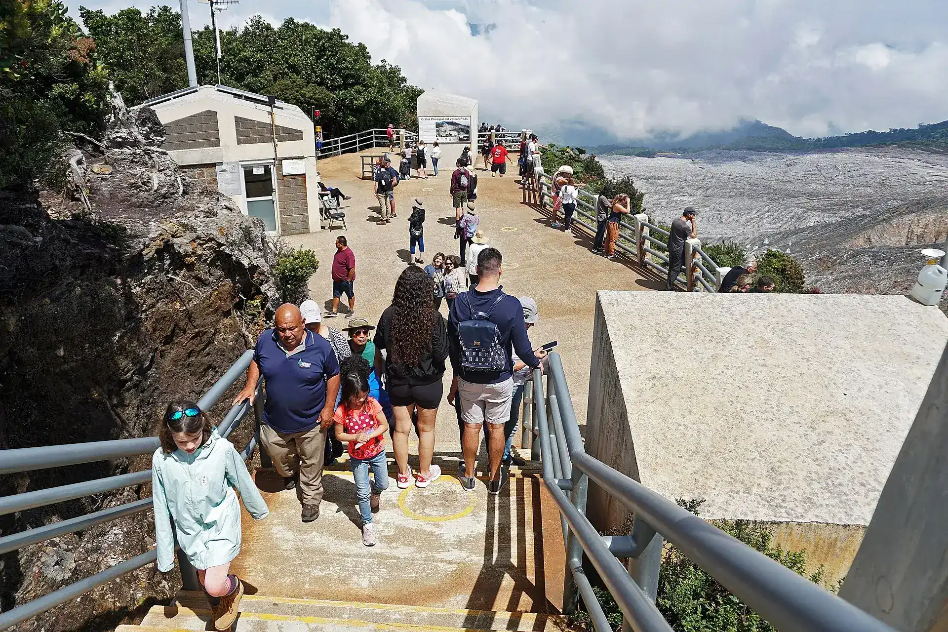 Visitors on the viewing platform at the crater rim of Poás Volcano next to a concrete shelter, the gray crater slopes in the background (April 2023)