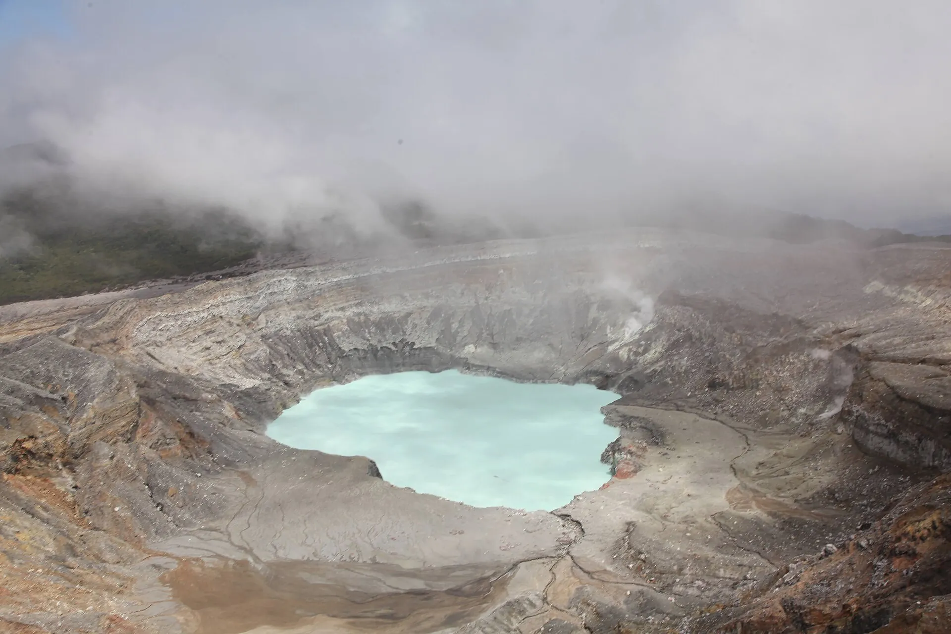View from the rim into the main crater of Poás Volcano: gray ash slopes, a milky turquoise acid lake, clouds drifting over the edge
