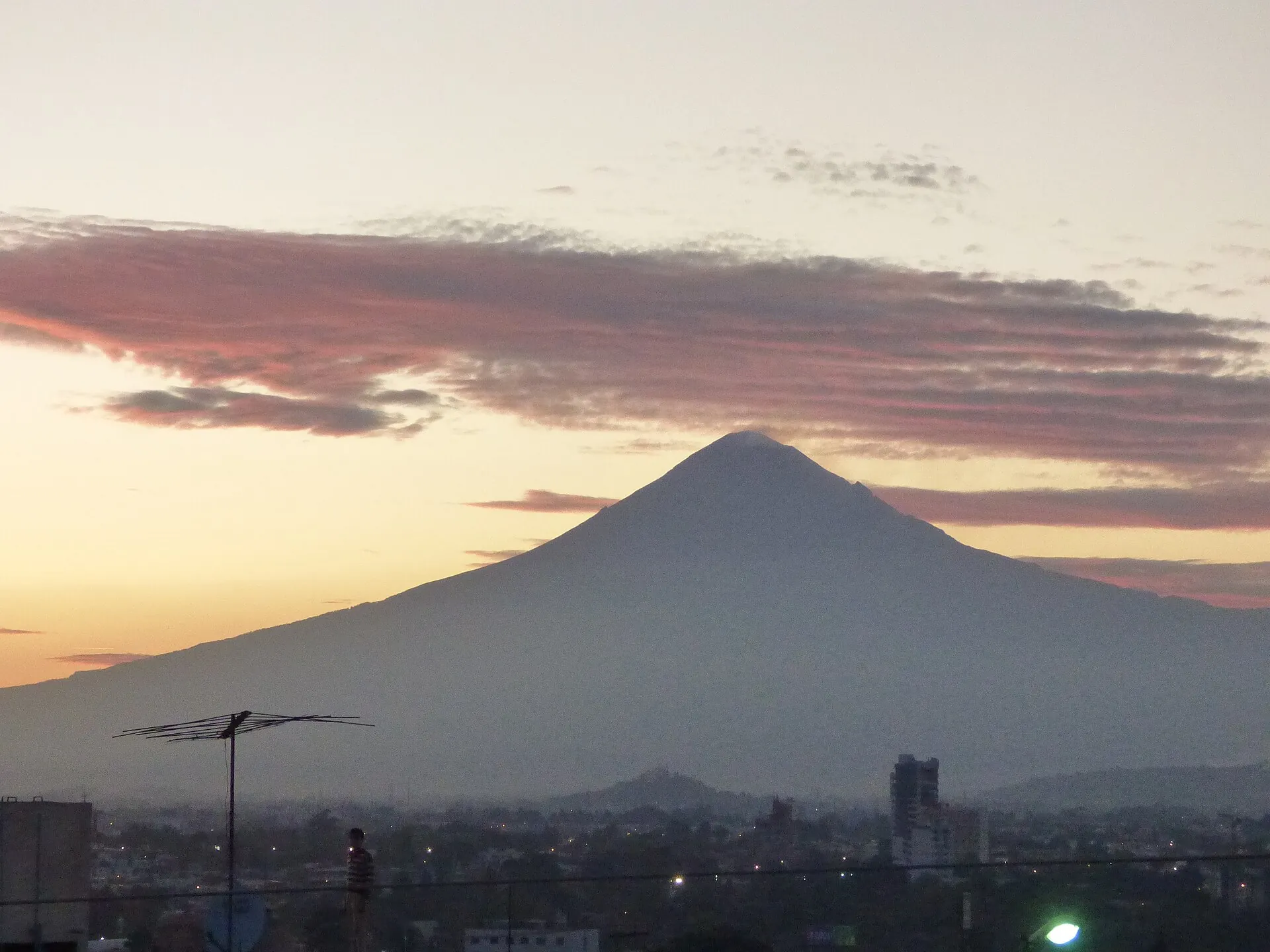 Der Popocatépetl als Silhouette vor orangefarbenem Abendhimmel über den Dächern von Puebla, im Vordergrund eine Palme