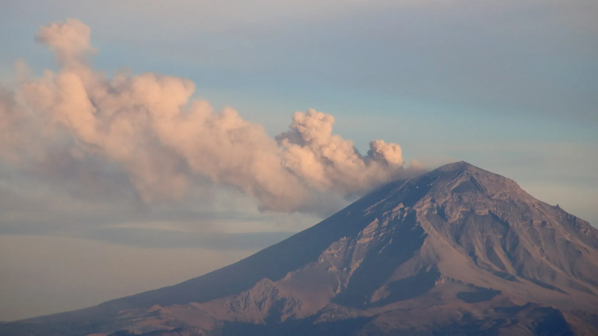 Der Popocatépetl stößt eine dichte, rosa-graue Aschewolke aus, die über den Kegel nach links zieht; Teleaufnahme aus Mexiko-Stadt