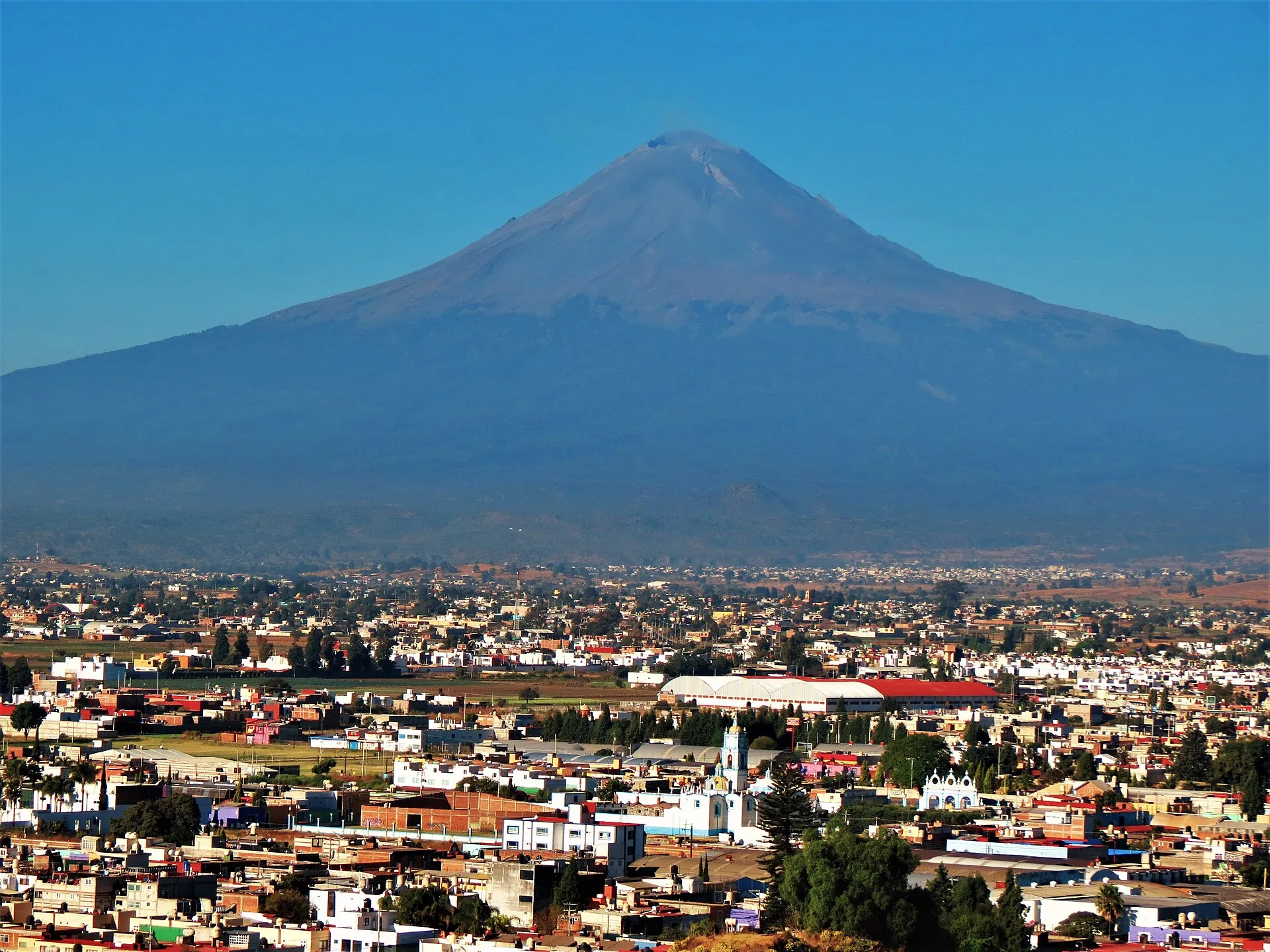 Der Popocatépetl über der Stadt Cholula bei Puebla: dunkler Kegel mit heller Fahne über dem Krater, davor Häuser und eine Kirche