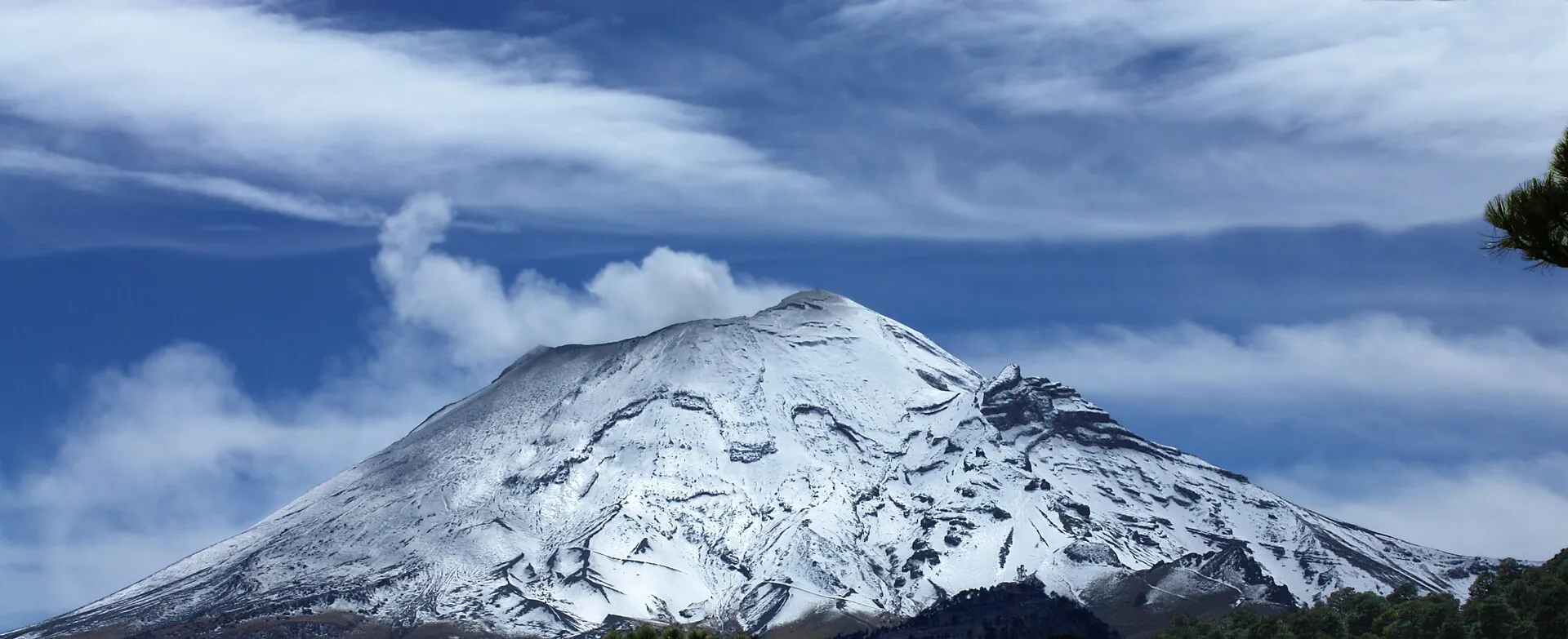 Der Popocatépetl vom Paso de Cortés aus: schneebedeckter Kegel mit weißer Dampffahne unter blauem Himmel mit Schleierwolken