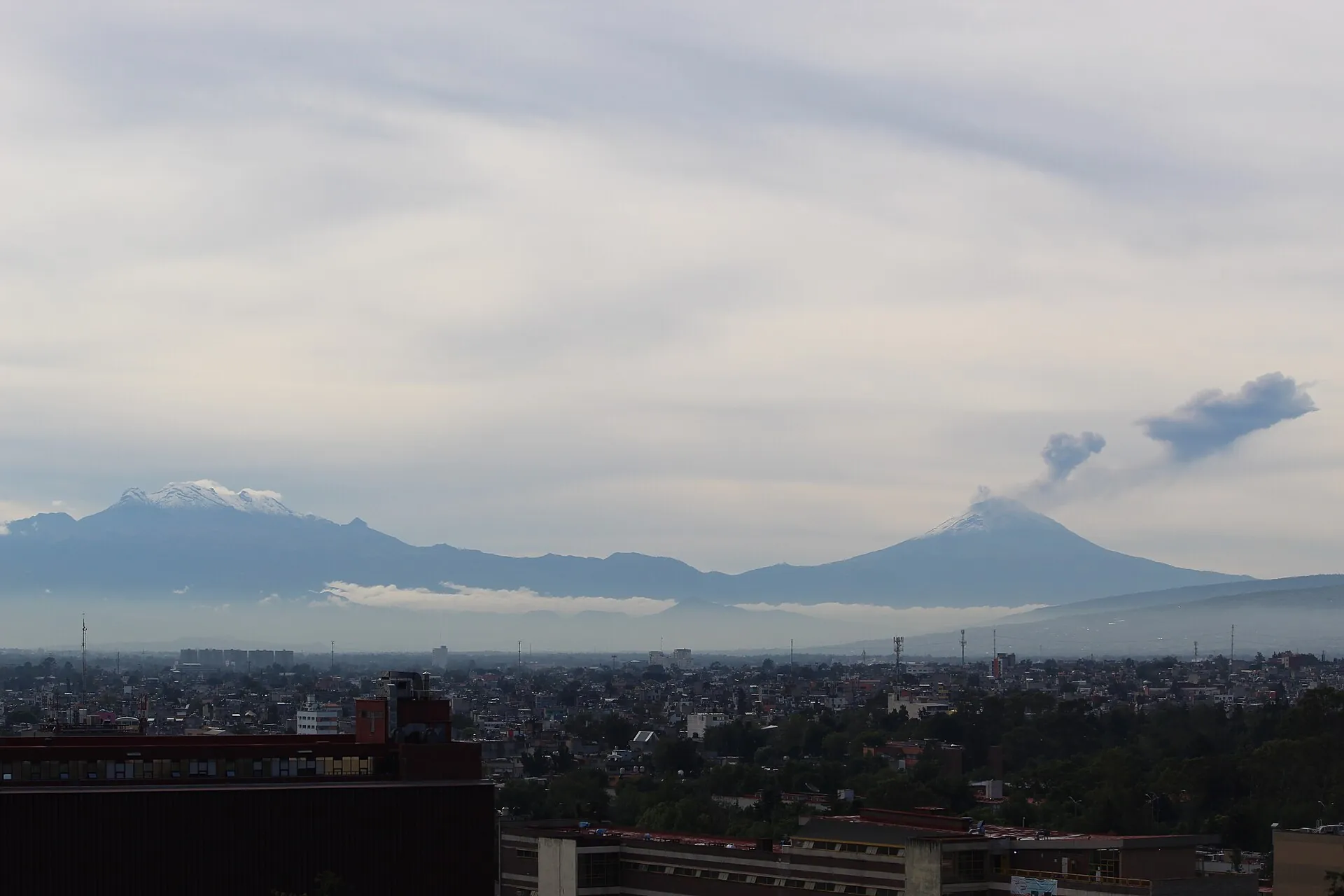 Popocatépetl und Iztaccíhuatl über den Dächern von Mexiko-Stadt: zwei schneebedeckte Gipfel am Horizont, über dem rechten eine Dampffahne