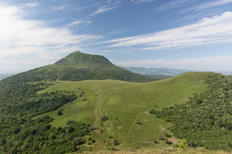 Blick vom Puy de Dôme über die Chaîne des Puys: grasbewachsene Kegel und Krater in einer Reihe unter blauem Himmel