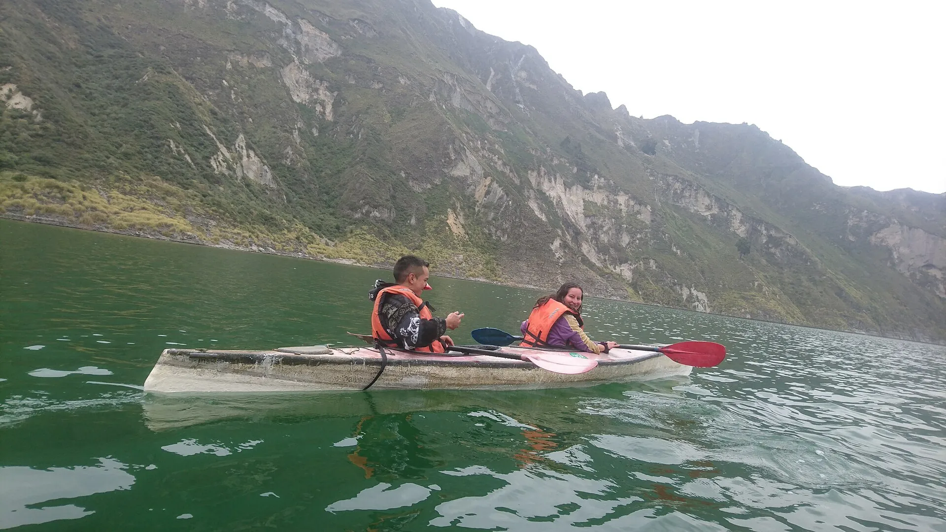 Two people in life jackets paddling a kayak across the green Laguna Quilotoa, the crater wall behind