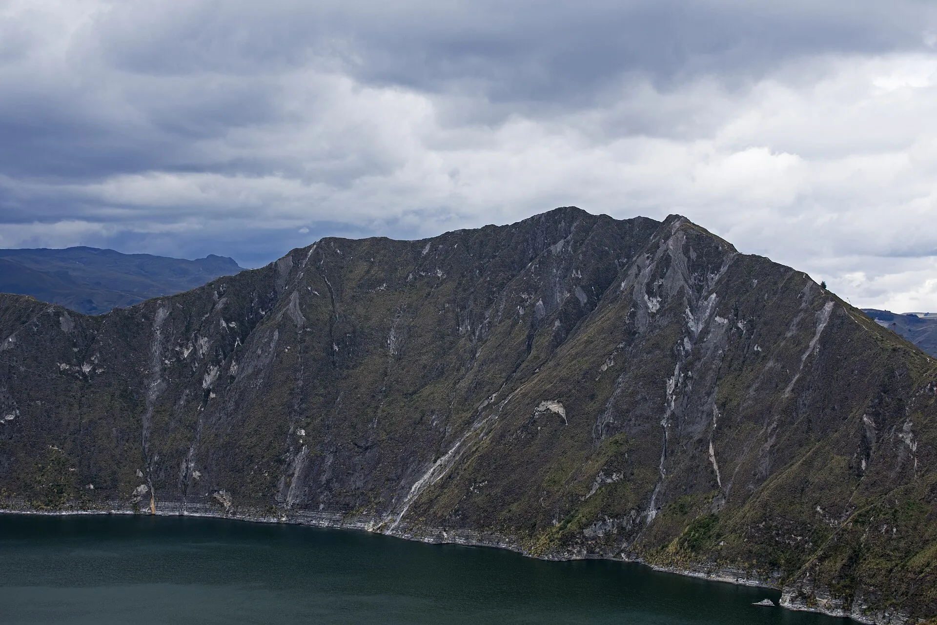 The steep, furrowed crater wall of Quilotoa dropping to the green lake, gray clouds above