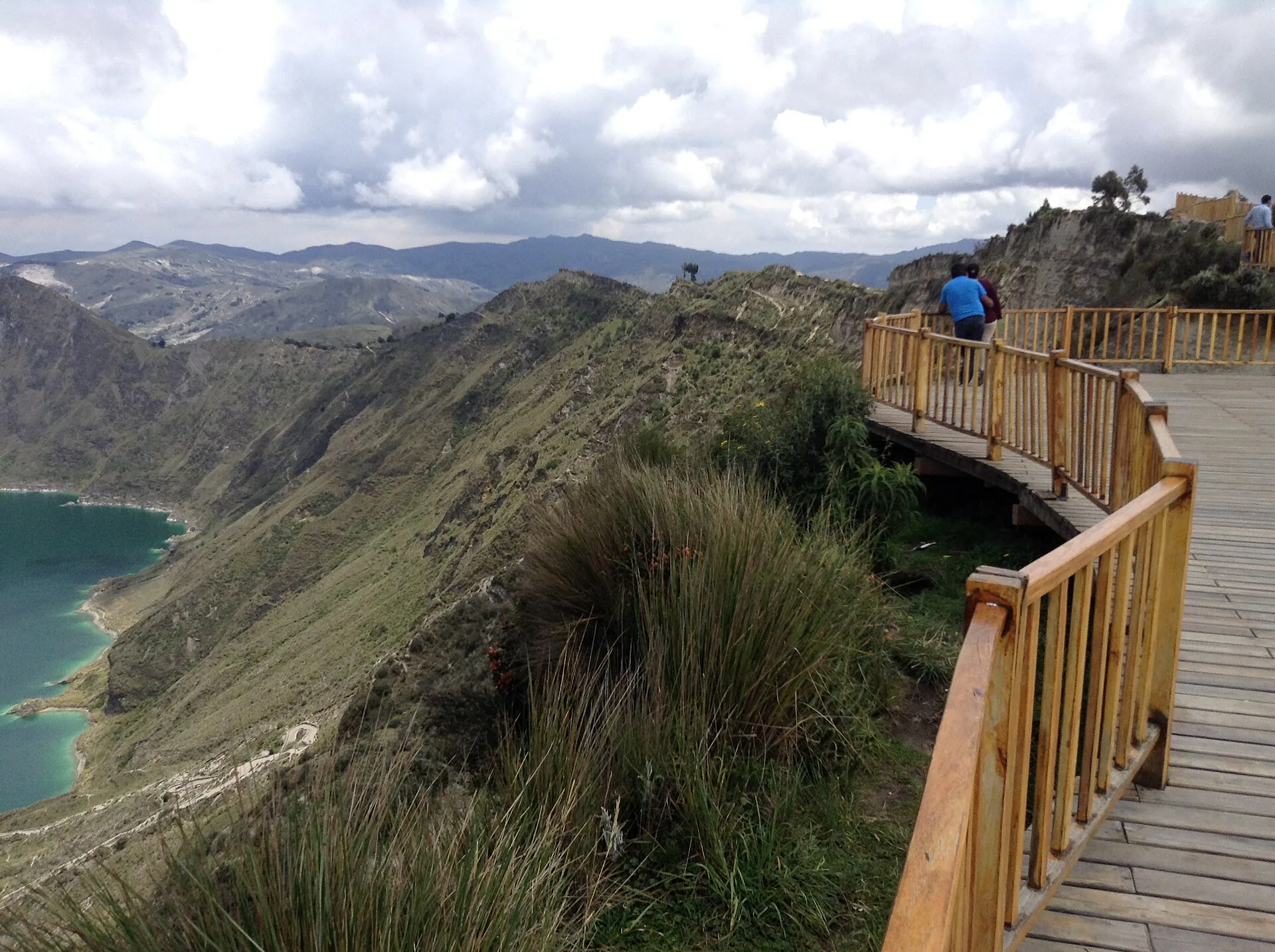 A curved wooden walkway with railing at a viewpoint on the Quilotoa crater rim, the lagoon below