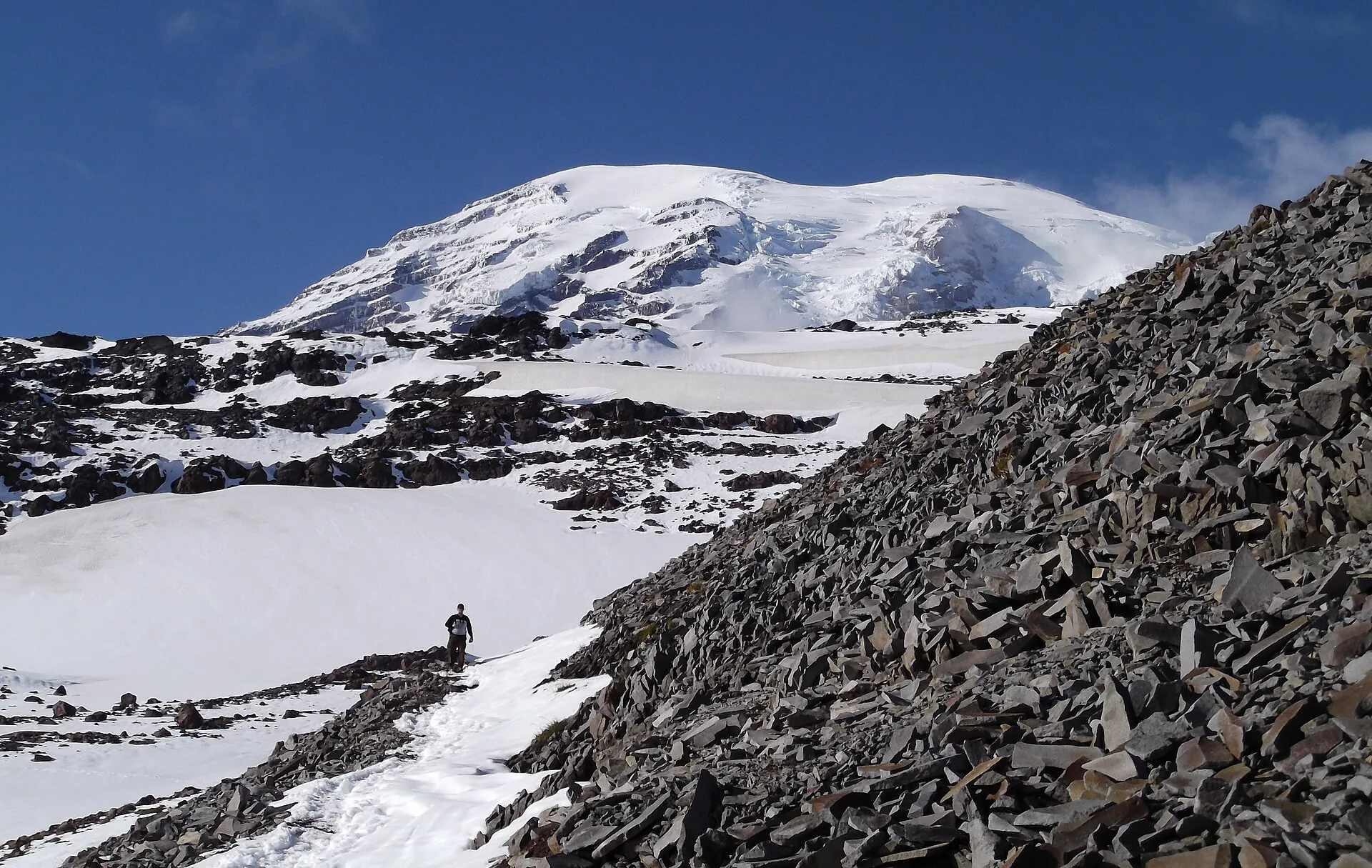 Ein einzelner Wanderer auf einem Schneefeld am Skyline Trail im Mount-Rainier-Nationalpark, darüber die vergletscherte Gipfelkuppe