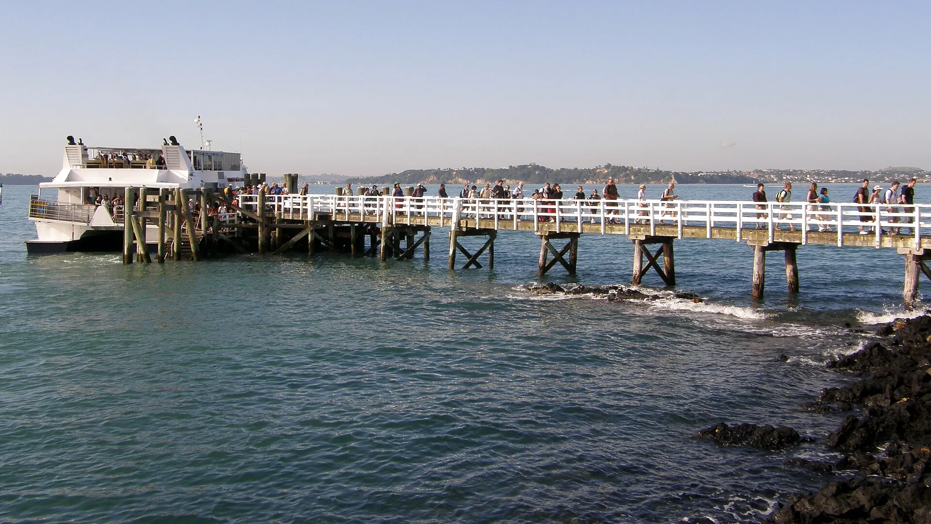 The ferry alongside Rangitoto Wharf, visitors walking down the long white wooden jetty to the island, with the Auckland coast behind