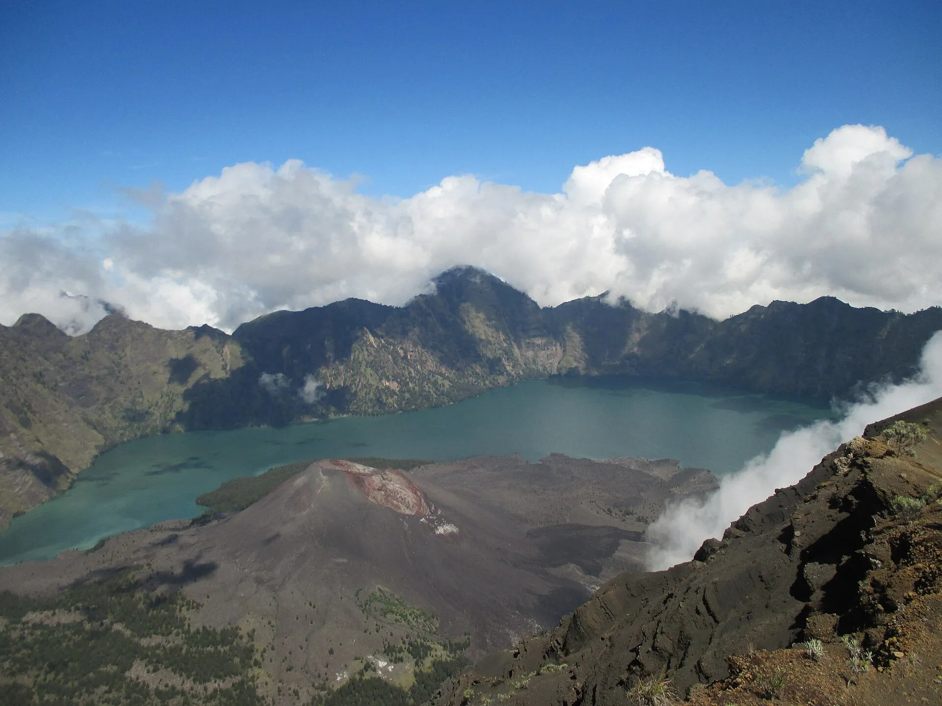 View from the crater rim of Mount Rinjani onto the crater lake Segara Anak and the dark cone of Barujari, with clouds above the caldera wall