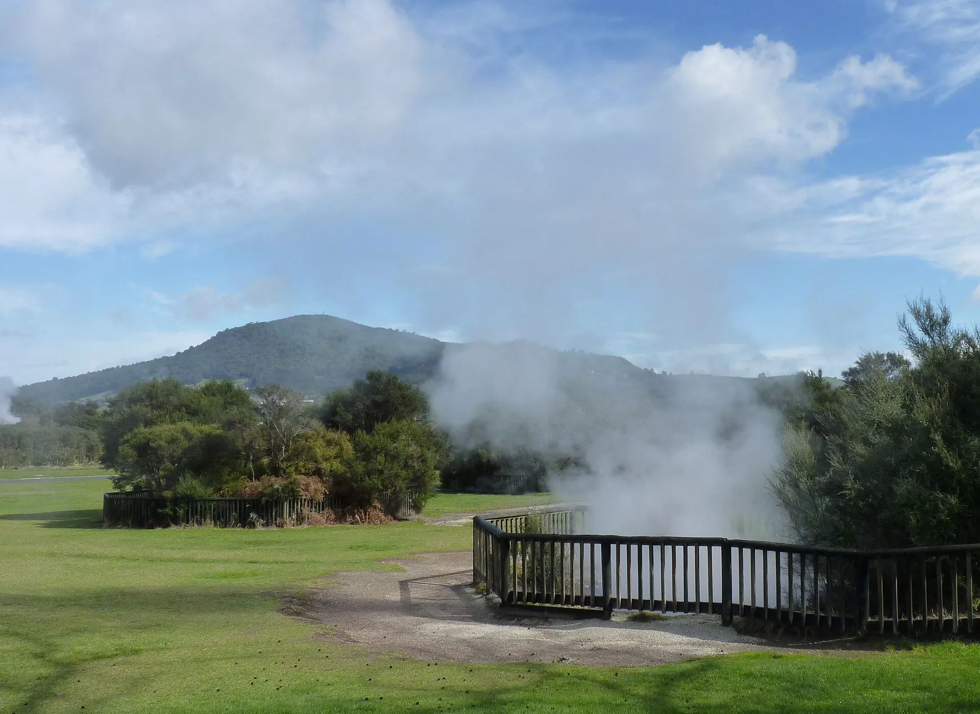 Kuirau Park in the middle of Rotorua: steam drifting across a lawn behind a wooden railing, with bush-covered hills beyond