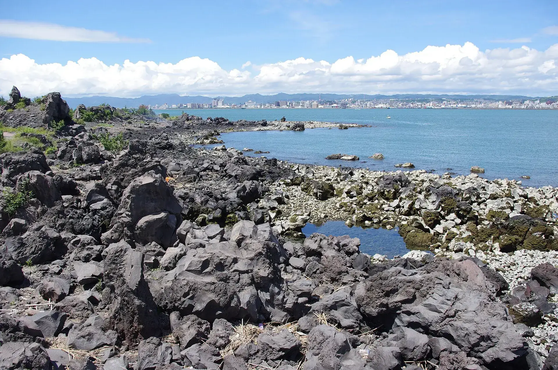 Black, sharp-edged lava blocks on the shore of Sakurajima, with Kinko Bay and the high-rises of Kagoshima behind