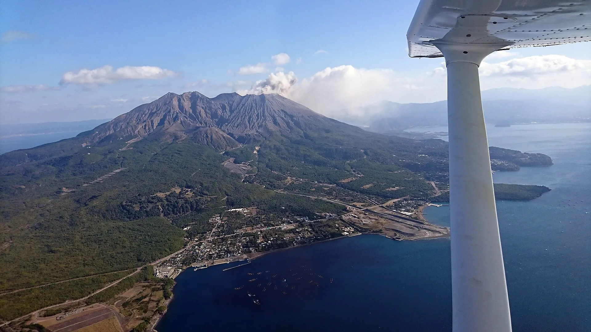 Aerial view from a light aircraft of Sakurajima with an ash plume above the summit, coastal settlements and Kinko Bay below