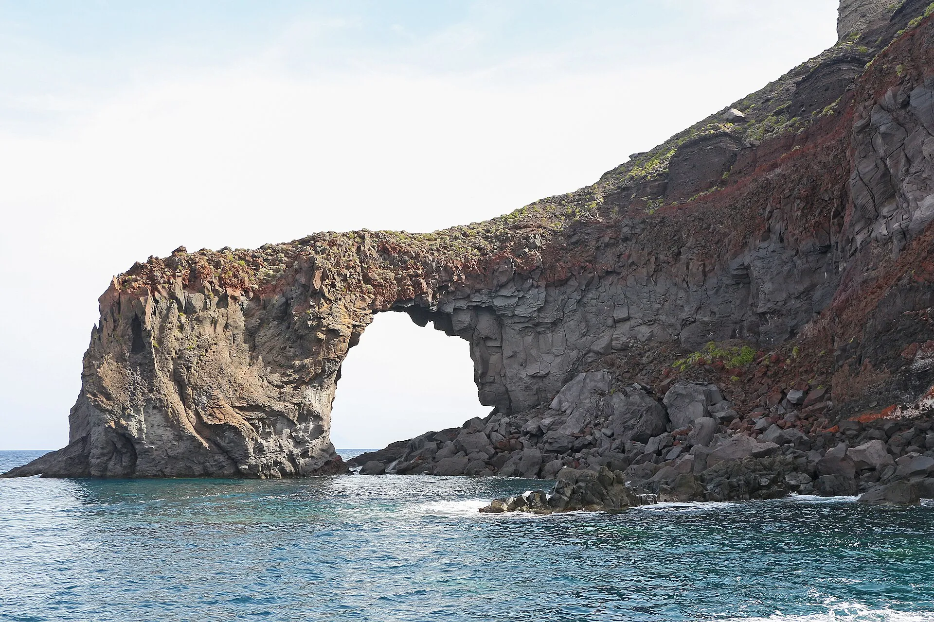The rock arch Punta del Perciato on the coast of Salina, dark volcanic rock above turquoise water