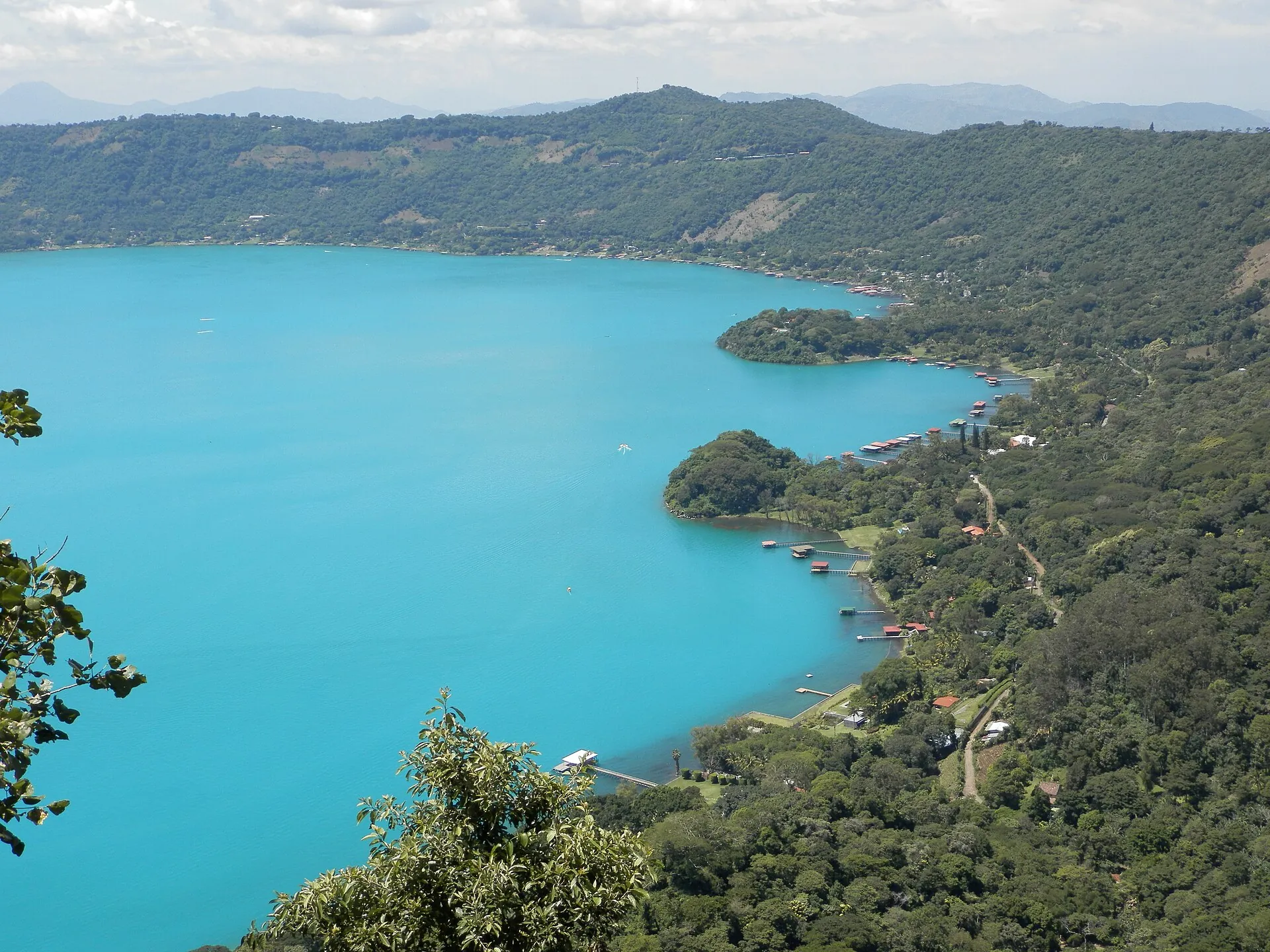 Lake Coatepeque in bright turquoise, houses and jetties on the shore, forested crater slopes behind