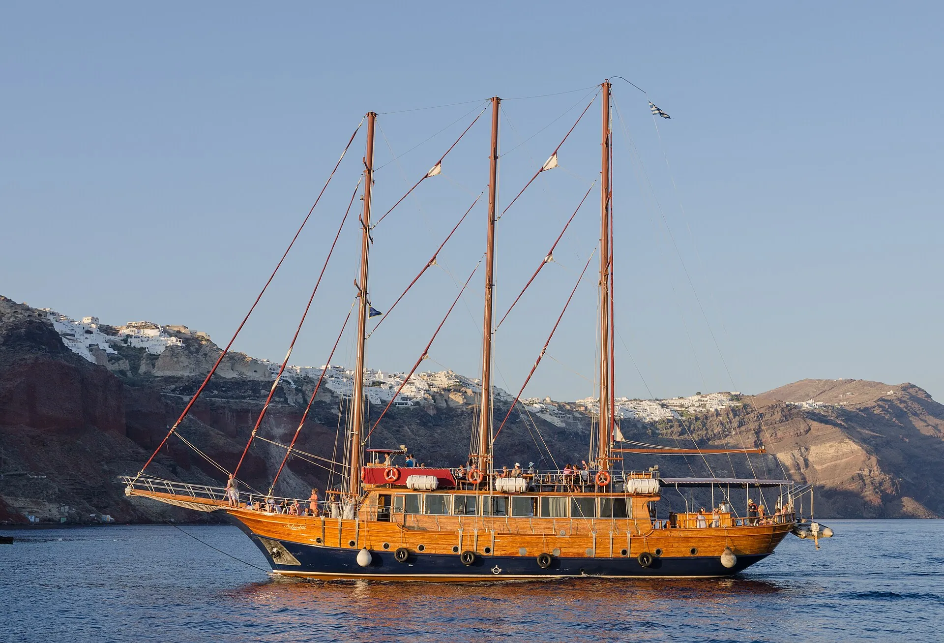 A three-masted wooden sailing ship with an orange hull lies off the caldera wall of Santorini