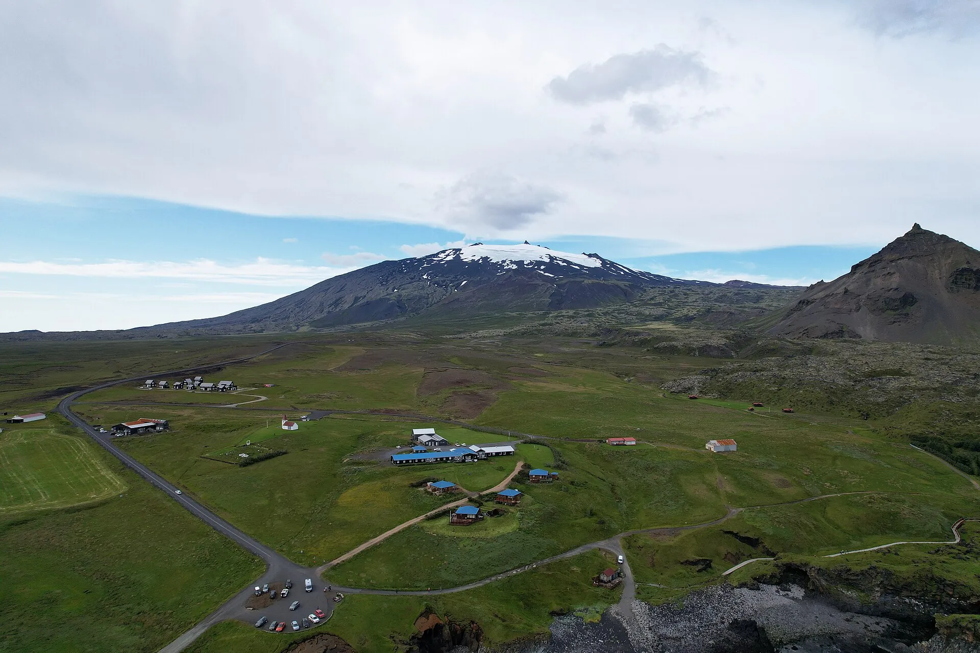 Luftaufnahme von Arnarstapi: das Fischerdorf an der Küste, dahinter der Berg Stapafell und der verschneite Snæfellsjökull