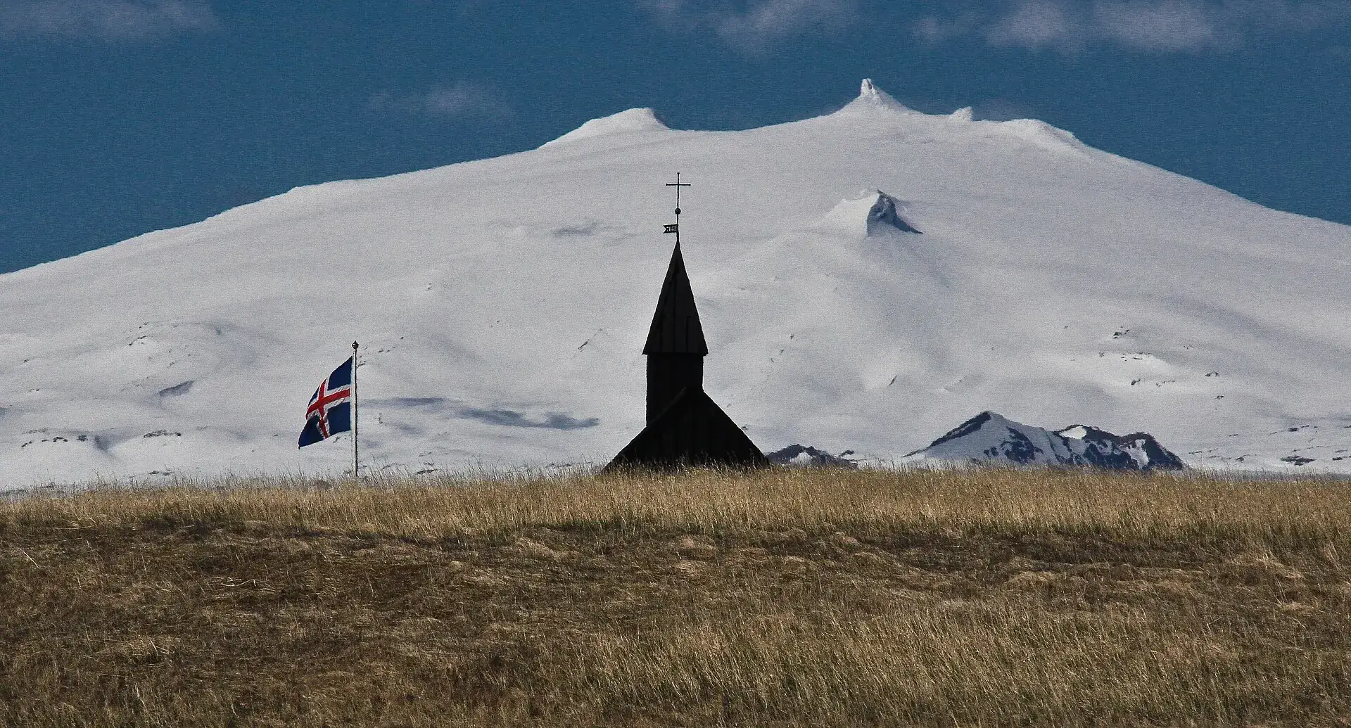 Die schwarze Kirche von Búðir vor dem verschneiten Snæfellsjökull, isländische Flagge im Wind