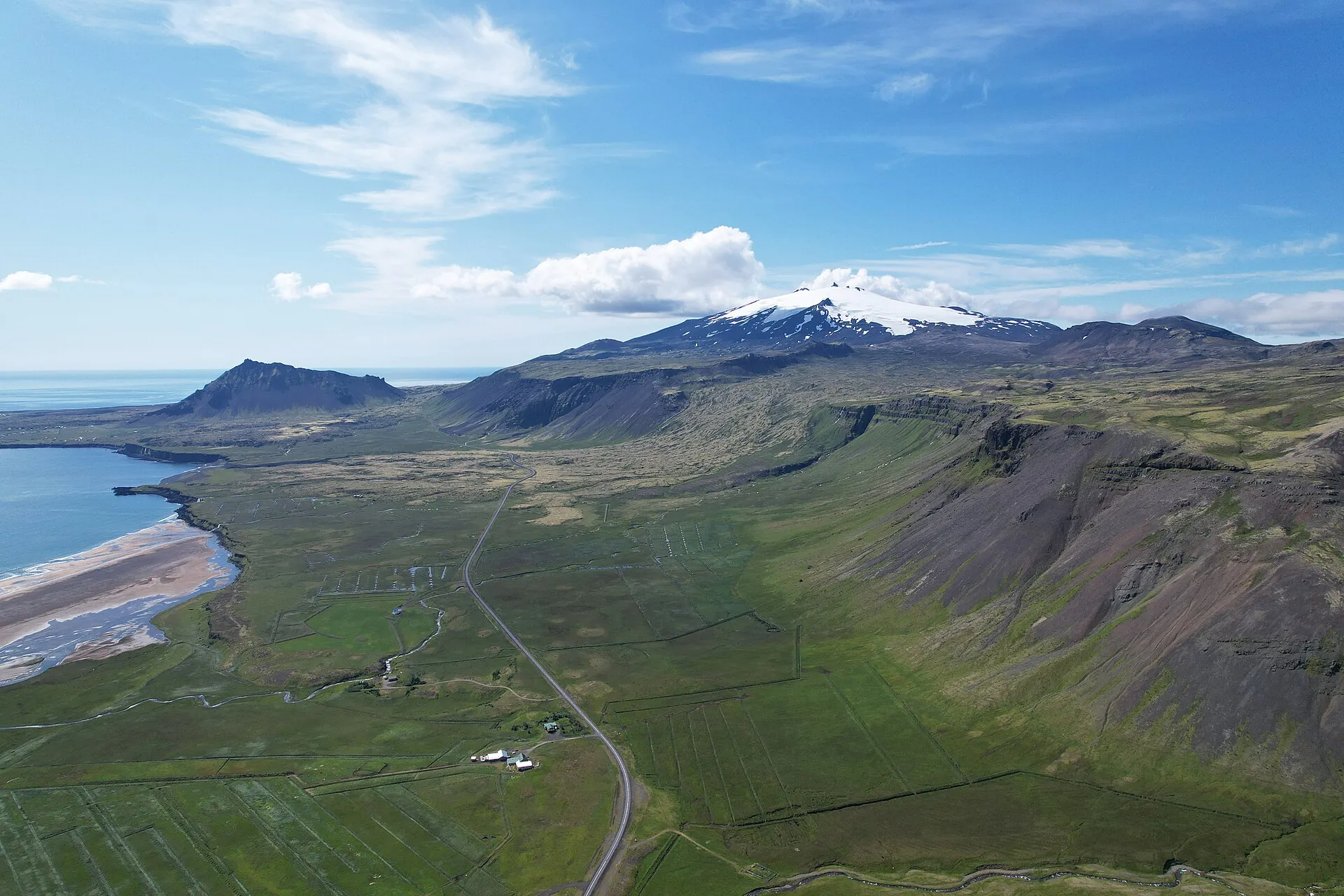 Luftaufnahme des Snæfellsjökull: der verschneite Vulkankegel über grünem Küstenland, eine Straße windet sich am Hang entlang